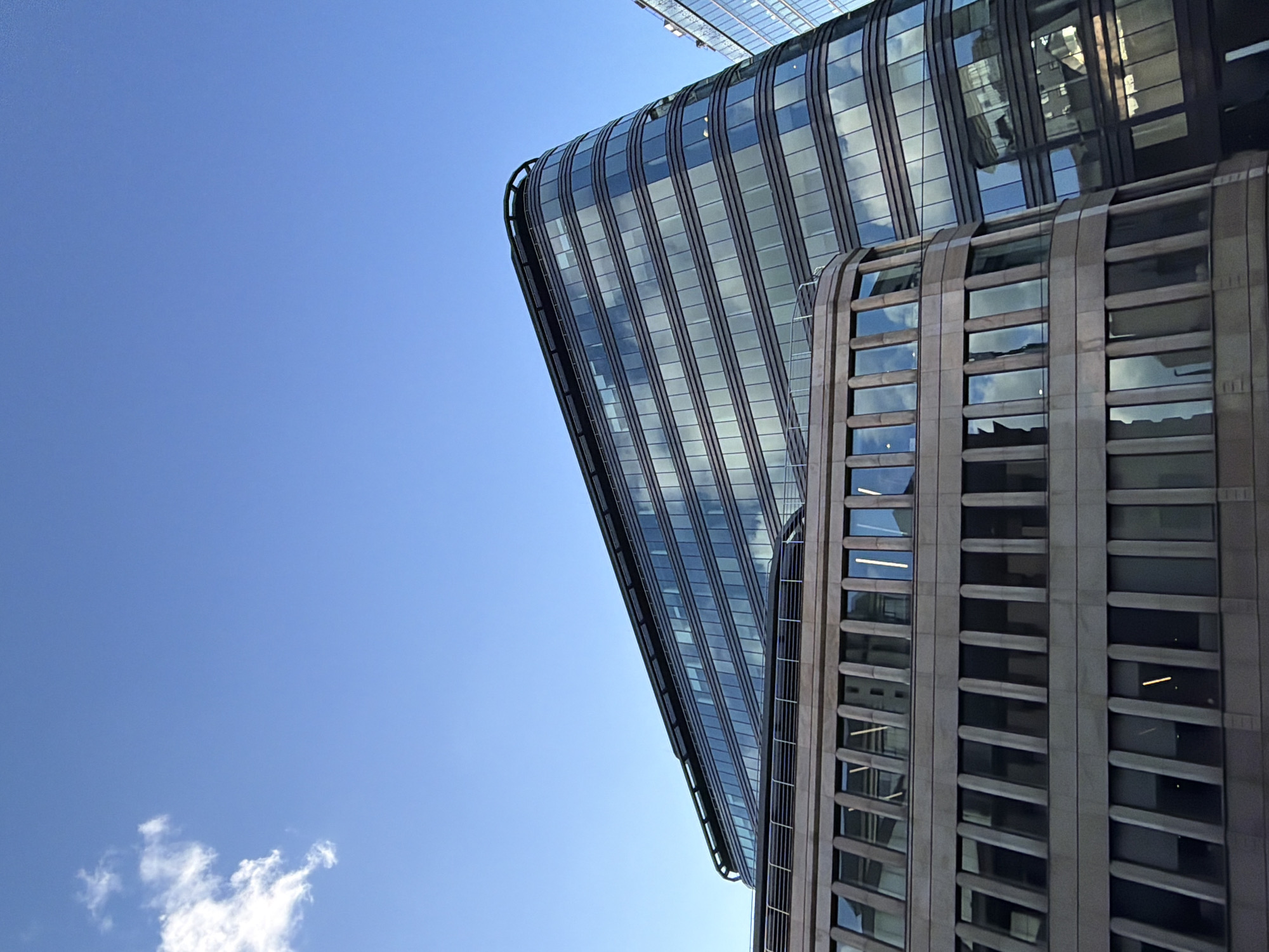 The image shows a low-angle view of two modern buildings against a clear blue sky with a few clouds at the bottom left corner. The building on the left is a glass skyscraper with rounded edges and visible steel supports. The building on the right is shorter and has a facade of stone or concrete with recessed windows. The image is rotated counter-clockwise.