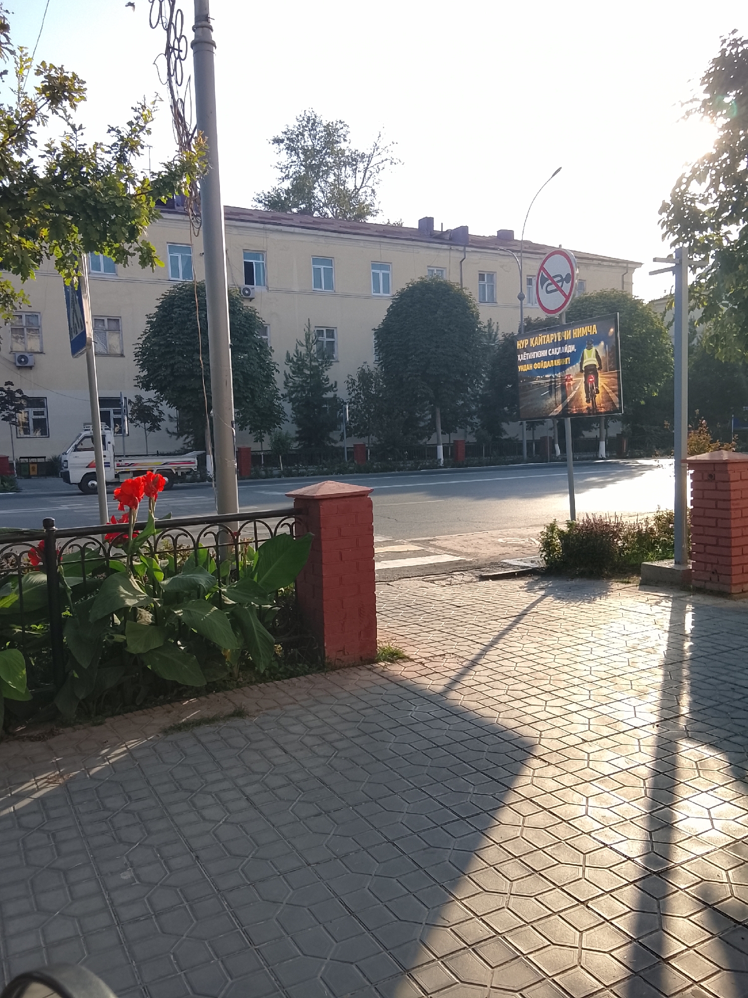 The image shows a street scene in Fergana City, Uzbekistan, on a sunny day. In the foreground, red canna lilies are in bloom behind a black metal fence. The sidewalk is paved with grey interlocking stones, and a shadow of a tree or structure is cast across it. To the left, a light-colored building with multiple windows stands behind a street lamp. Across the street, a white truck is parked. A large billboard displays an image of a person on a bicycle and text in a foreign language. In the background, more trees and buildings are visible.