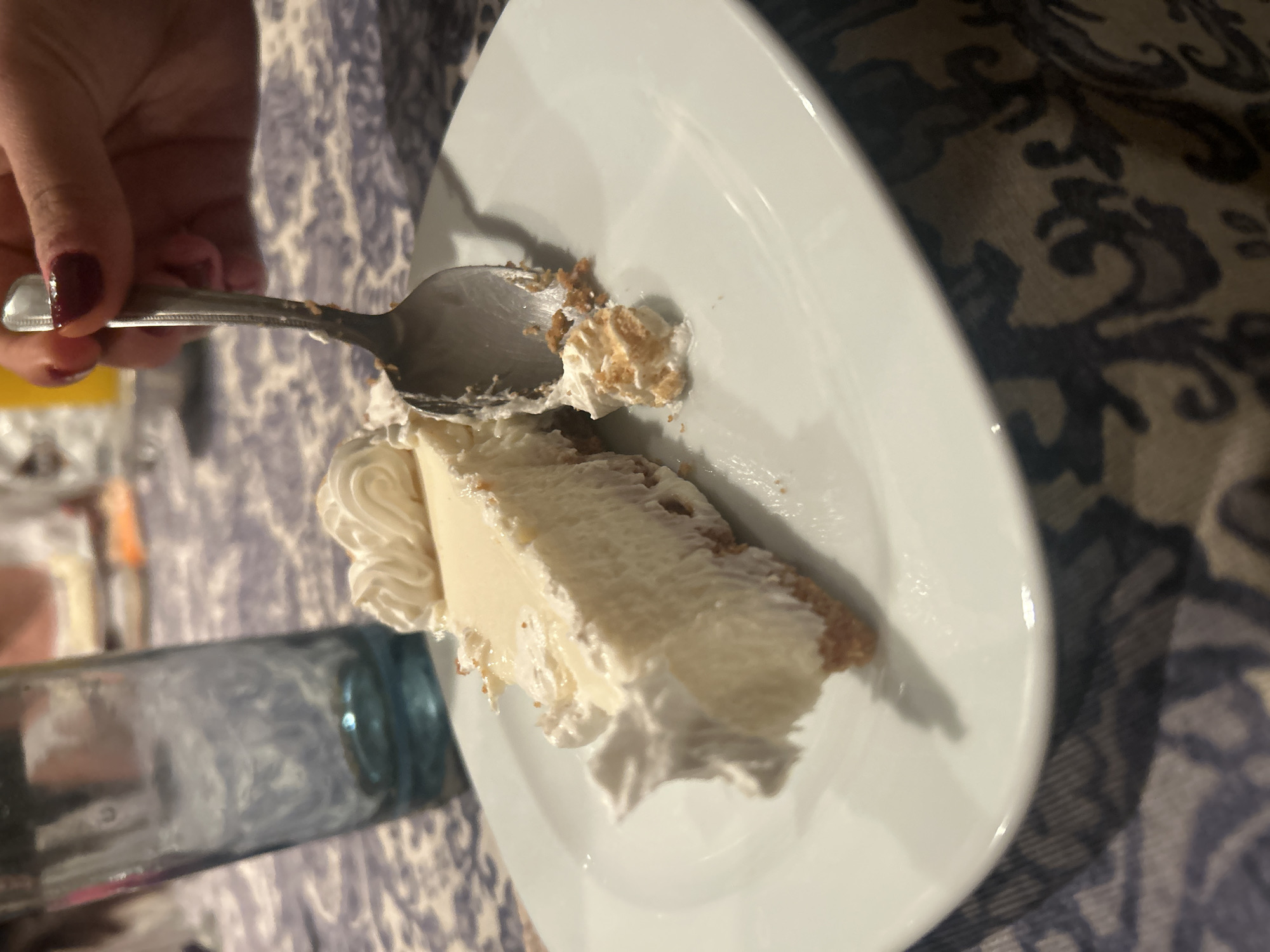 A close-up shot depicts a partially eaten slice of key lime pie on a white plate. A hand holds a silver spoon with a portion of the pie on it. Next to the plate is a partially visible blue glass. The background features a tablecloth with a patterned design. The location is Unknown, Unknown.