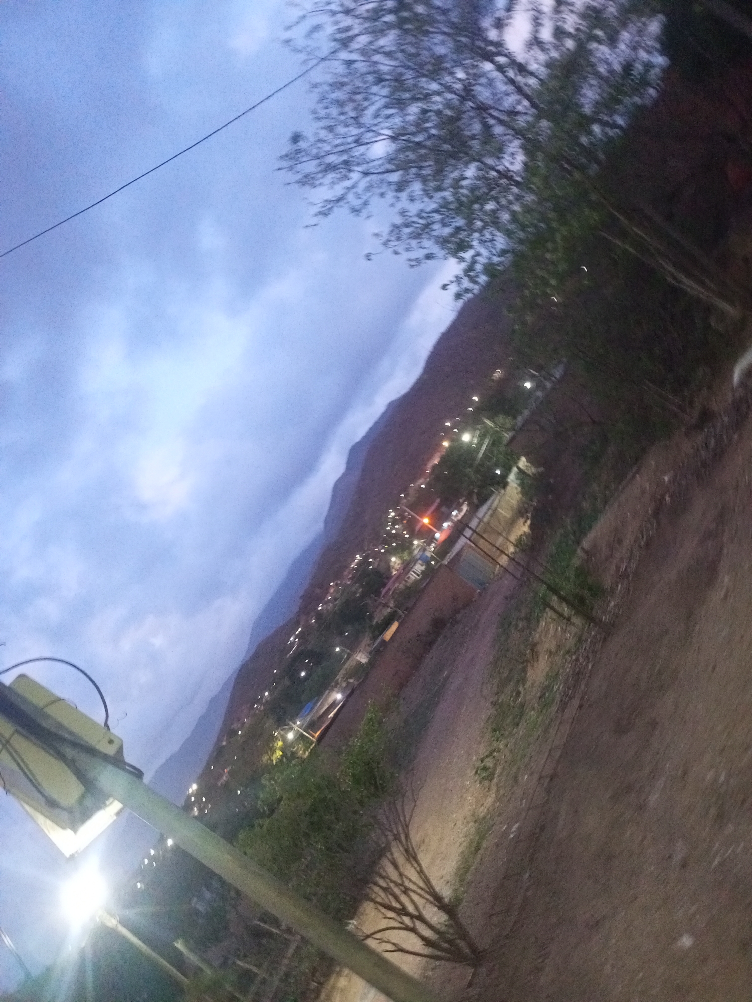 The image captures a twilight scene in the city of La Tagua, Venezuela, with a focus on a utility pole in the foreground. The sky is a blend of deep blue and lighter grays, with visible clouds, suggesting the transition from day to night. A thin black wire stretches diagonally across the upper left portion of the frame. To the right, a tree with sparse leaves is visible, with its branches blurred due to motion.
In the background, a mountainous landscape dominates. A town is nestled along the mountainside, illuminated by numerous lights that appear as small, bright dots. These lights suggest a populated area with buildings and streetlights. The terrain in the foreground is rough and unpaved, with a dirt path winding through it. Vegetation, including grass and bushes, is sparsely distributed along the path and on the slopes. The utility pole in the foreground is illuminated by a bright light, casting a strong glow. Another light fixture is visible on a secondary pole further down the path. The overall atmosphere is one of a rural or semi-rural setting at dusk.