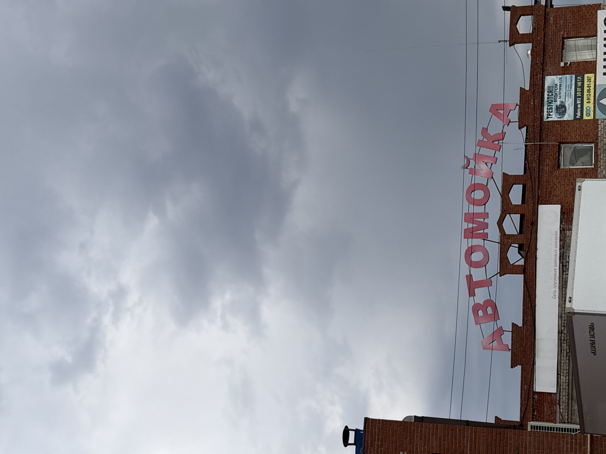 The image shows a cloudy sky above a brick building in Verkh-Sosnova, Russia. A large red sign reading "АВТОМОЙКА" (Car Wash) is attached to the building. Various signs and advertisements are visible, including one that reads "ТРЕБУЮТСЯ!!!" (REQUIRED!!!) for drivers. Another advertisement includes the text "«КАЗАЧЬЯ ЗАСТАВА» Сеть грузовых шинных центров" (Cossack Outpost - Chain of truck tire centers). A phone number is visible on one of the signs.