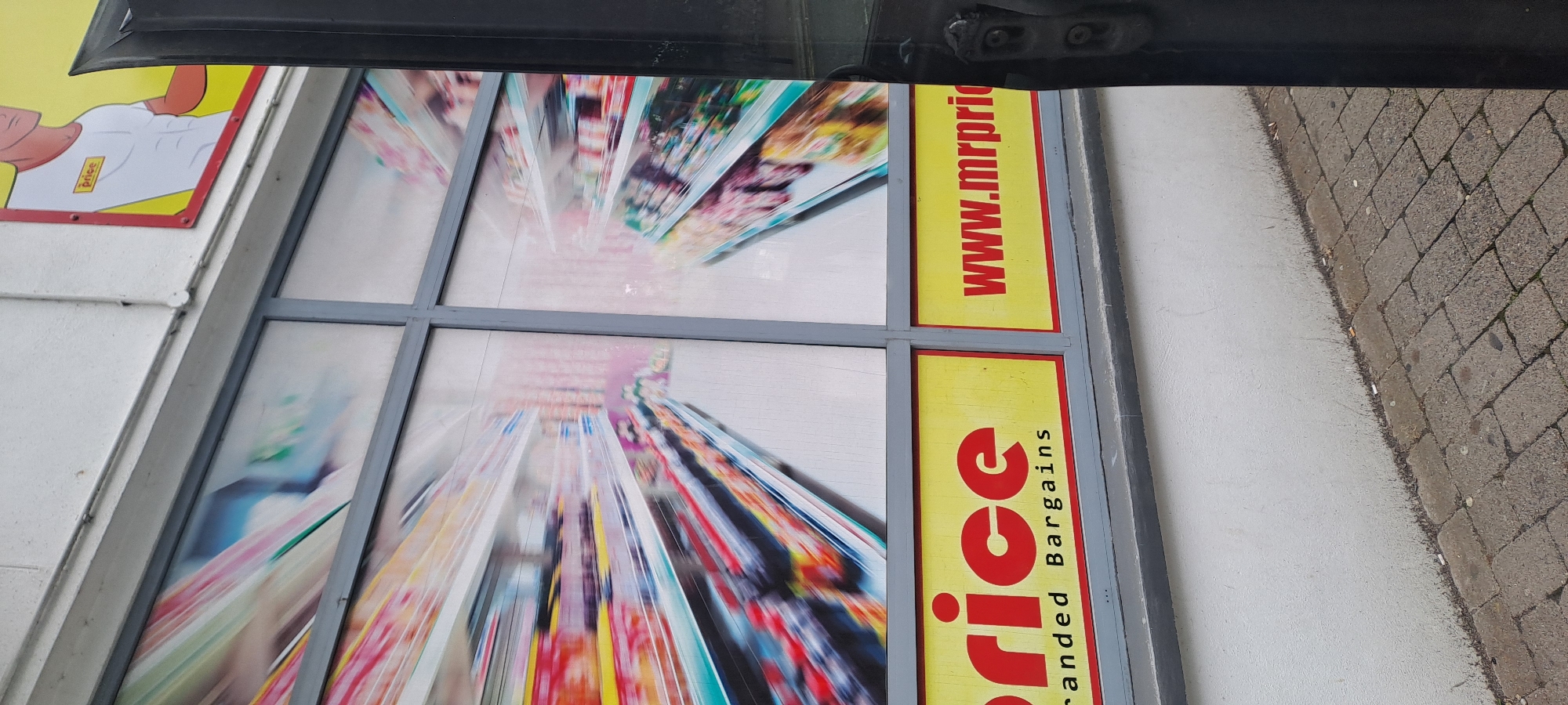 The image shows a storefront with large windows featuring blurry images of supermarket aisles. A yellow sign with red text reads "price" and "Branded Bargains." Another yellow sign with red text to the right reads "www.mrprice." The store appears to be Mr. Price, a discount retailer. The location is identified as Castlebar, Ireland. A black doorframe is visible on the right side of the image, partially obscuring the storefront. The building's exterior wall is white, and a paved sidewalk is in front of the store. The overall scene suggests a retail environment with a focus on low prices.