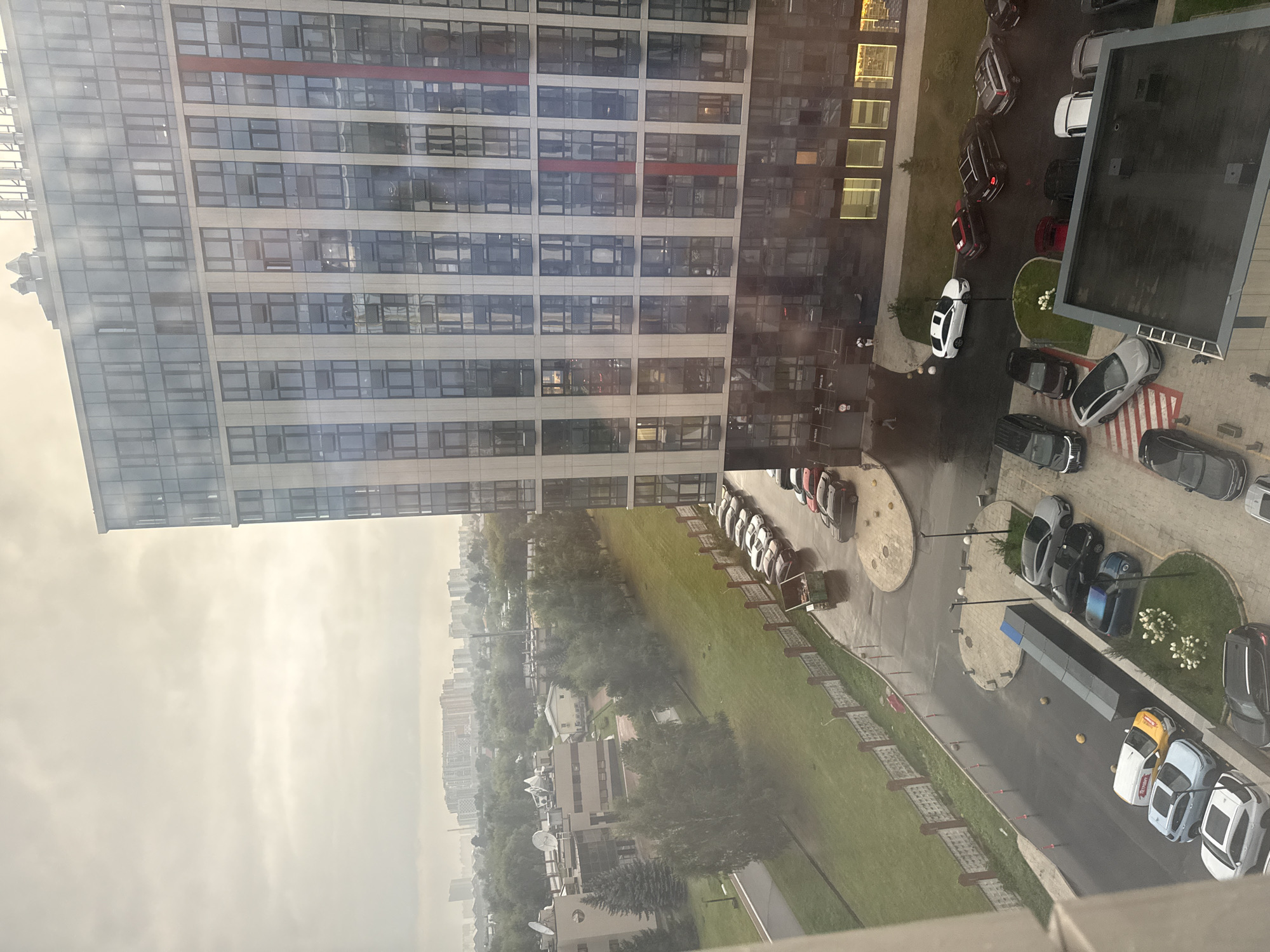 The image, captured from an elevated perspective, depicts a contemporary urban landscape under overcast skies, with a modern multi-story building prominently featured on the right. This building is characterized by its facade of numerous glass windows and vertical panels, creating a reflective surface. In the foreground and midground, a paved area with parked vehicles is visible, including various models of cars and a white utility van. Some of these vehicles are parked in designated spots, while others are situated along a roadside. A red and white striped marking is present on the pavement near some parked cars. A low, decorative fence runs along a green, grassy area adjacent to the road, bordering a sidewalk. Further into the distance, a wider cityscape unfolds, with more buildings and trees visible under a hazy sky. A dumpster is located near the fence line. The ground in the paved areas appears wet, suggesting recent rain. The overall scene is one of a residential or mixed-use urban environment.
