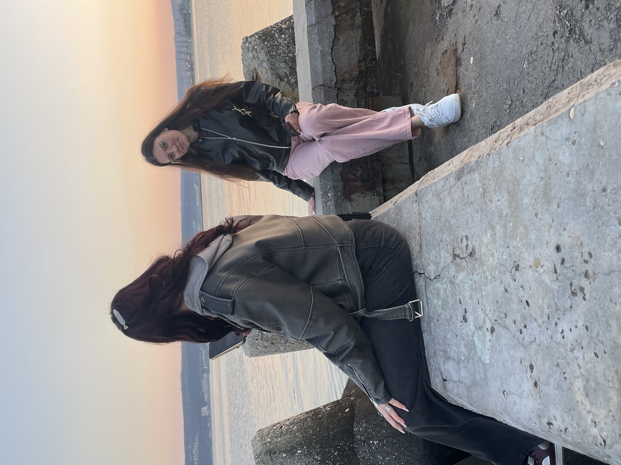 Two young women are positioned on a concrete structure with a body of water and city skyline in the background, indicative of a coastal location. The woman at the top is sitting and wearing a black leather jacket, pink pants, and white sneakers. She is looking at the camera and appears to be smiling. The second woman below her is wearing a gray hooded jacket, black jeans, and dark-colored sneakers. She is looking down and appears to be using her cell phone. The structure they are on is cracked and weathered concrete. The scene is set during the day with a sunset or sunrise visible in the sky. Based on the urban landscape and coastal setting, the location is most likely Varna, Bulgaria.