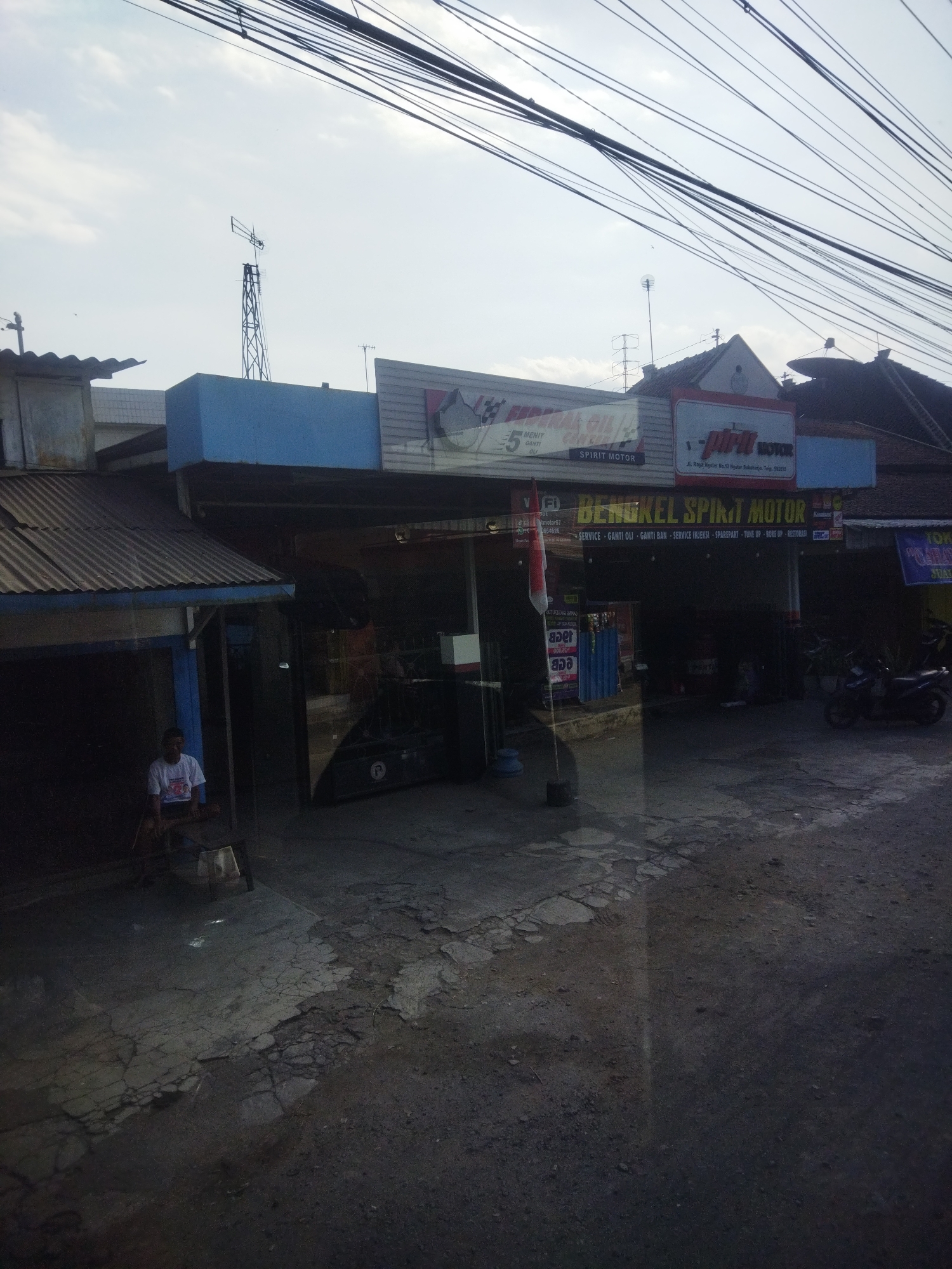The image depicts a street view in Nguter, Indonesia, featuring a motorcycle repair shop called "BENGKEL SPIRIT MOTOR." The shop is located on a paved street with some sections showing wear and tear. Overhead, a complex network of electrical wires crisscrosses the sky, obscuring a portion of the buildings and sky.
In front of the shop, a person wearing a white t-shirt and shorts is seated on the ground, facing away from the camera. A motorcycle is parked on the right side of the image, near the shop's entrance. The shop itself has a dark facade, with signs indicating its services, including "SERVICE - GANTI OLI - GANTI BAN - SERVICE INJEKSI - SPAREPART - TUNE UP - BORFES."
To the left of the shop, a building with a corrugated metal roof is visible. Further to the left, a tall, lattice-like antenna structure stands against the sky. The overall atmosphere suggests a typical commercial street scene in a local Indonesian town. The lighting indicates it's likely daytime, possibly late afternoon given the shadows.