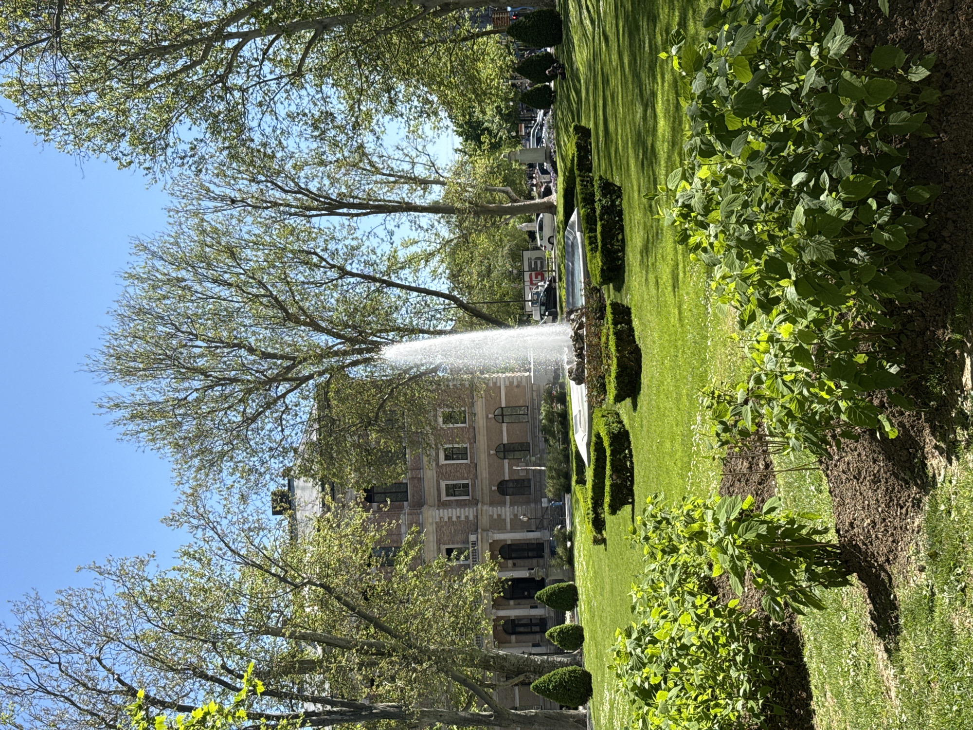 The image presents a park scene in the city of Zagreb, Croatia. A grand, multi-story building with arched windows and a light-colored facade is situated in the background. A tall water fountain shoots a plume of water in front of the building. The park is lush with green grass, bushes, and precisely trimmed hedges. Trees with budding leaves frame the scene. A few people are visible in the distance, near parked cars. The sky is a clear, bright blue, suggesting a sunny day.