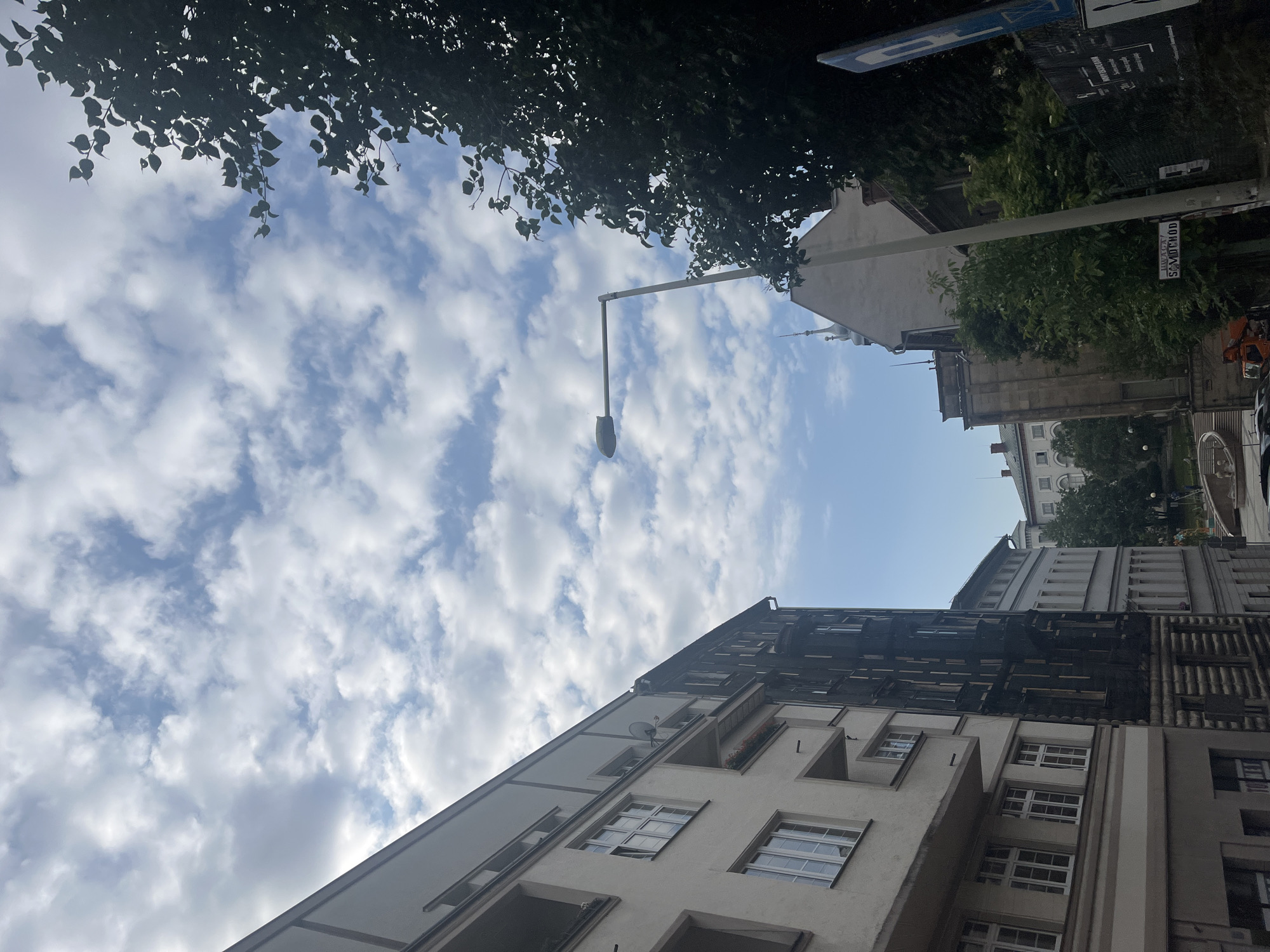 The image shows a street scene with buildings and a cloudy sky. On the left, there is a light-colored apartment building with multiple windows and balconies. To its right, a darker, older building appears to be undergoing renovation or has a facade covered in some material. Further down the street, more buildings are visible, including one with a pointed roof. A lamppost stands prominently in the center right of the frame, with a large tree providing shade on the far right. The sky is filled with scattered white and grey clouds against a blue background. Signs are visible on a fence in the foreground on the right. The scene appears to be in an urban environment.