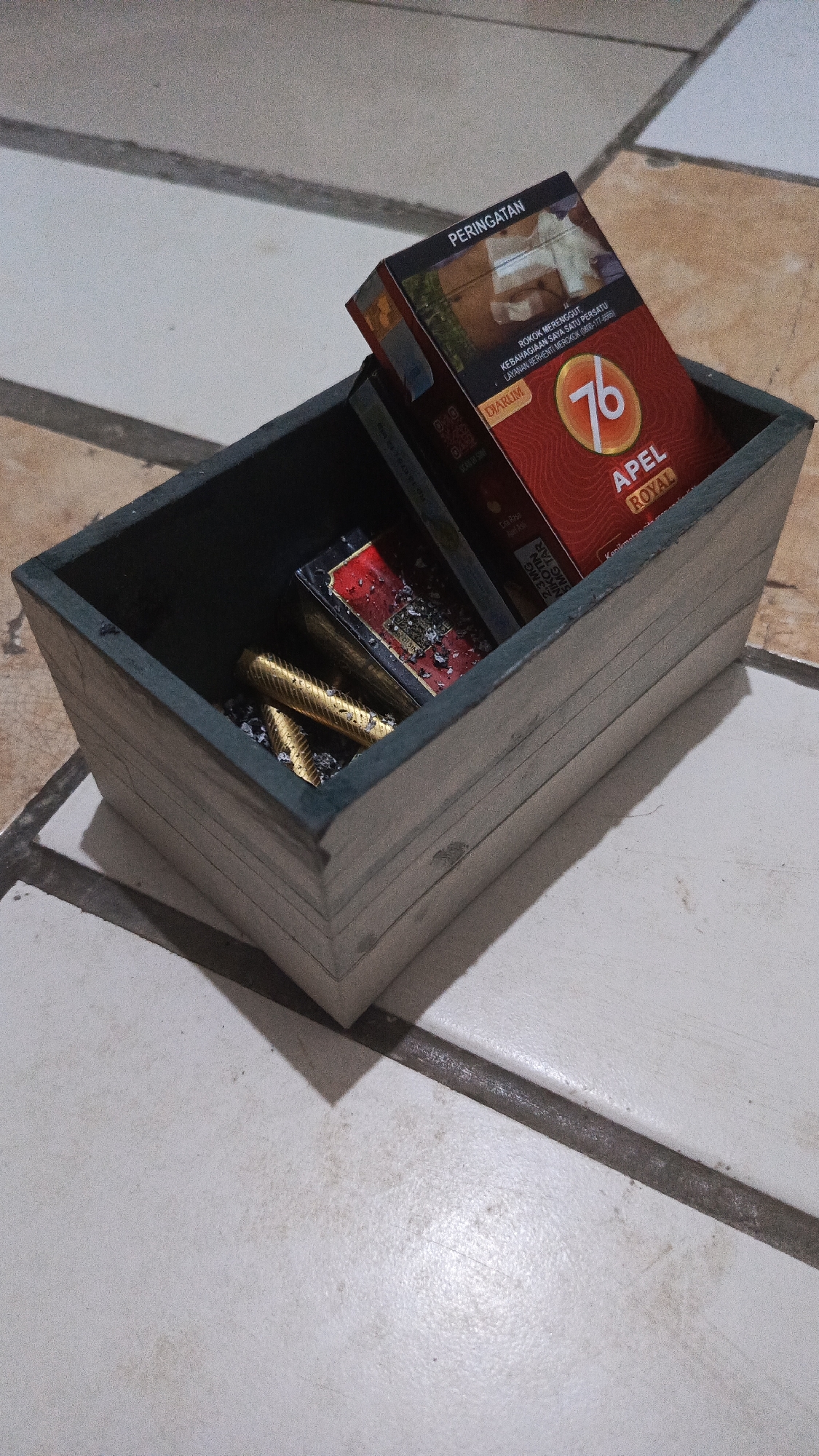 A wooden box, likely an ashtray, sits on a tiled surface. The box contains two packs of cigarettes, one with a red and gold design and the brand "76 APEL ROYAL" clearly visible. The other cigarette pack is partially obscured. Several gold-colored cigarette butts, possibly remnants of smoking, are also present in the box, along with scattered ash. The overall scene suggests a casual setting, possibly a home or an outdoor seating area in Tangerang, Indonesia. The tiling appears to be a common design found in many urban environments in the region.