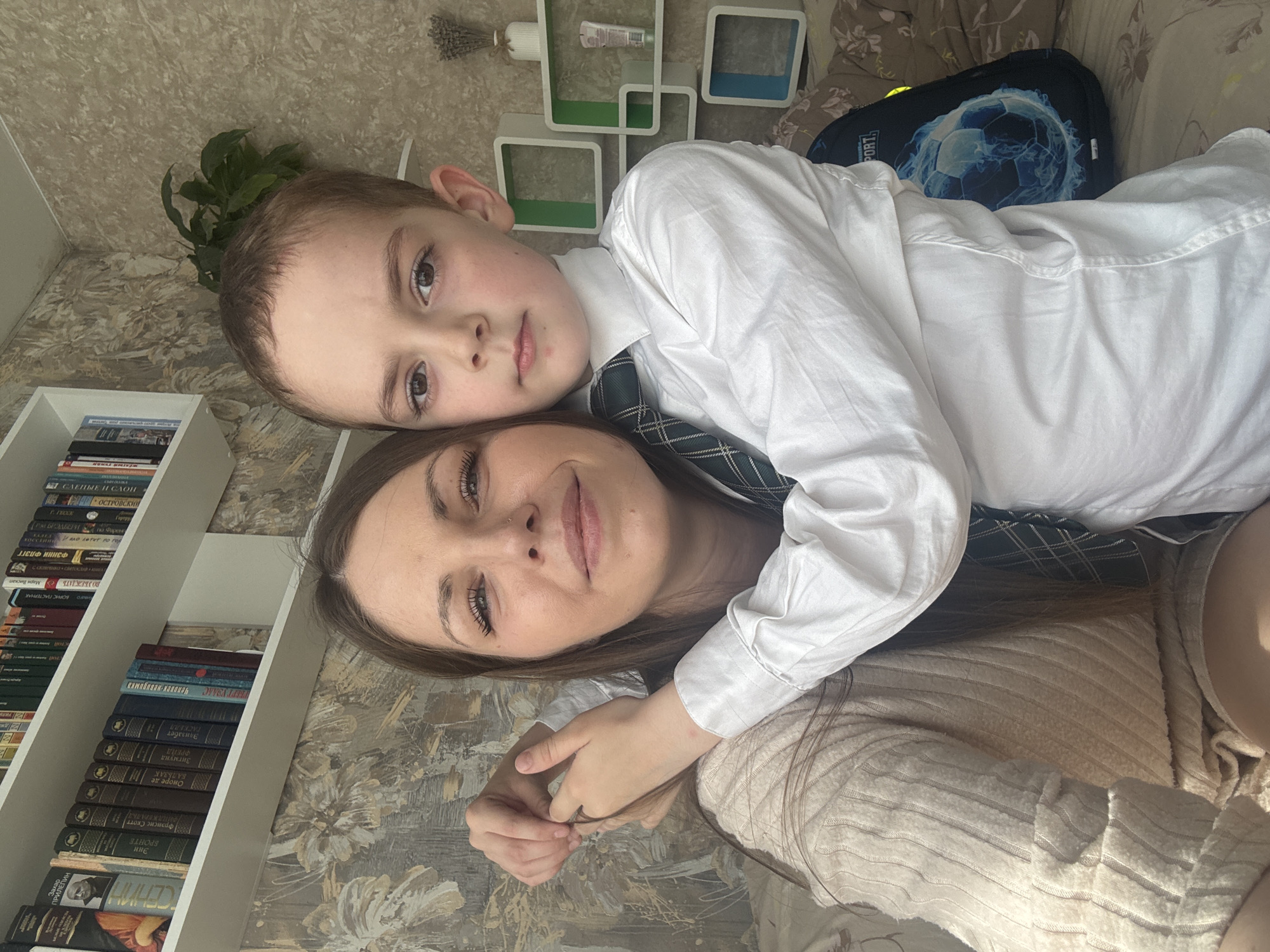 A woman with long brown hair is lying down, with a young boy lying on top of her. The boy is wearing a white shirt and a green and navy tie. They are both looking at the camera. The woman is wearing a beige sweater. Behind them is a wall with floral wallpaper, a white bookshelf filled with books, and some decorative white shelves. A blue and black backpack is on the right side of the frame. Based on the presence of Russian language books on the shelf, the location is likely in Russia, potentially in the city of Раздолье.