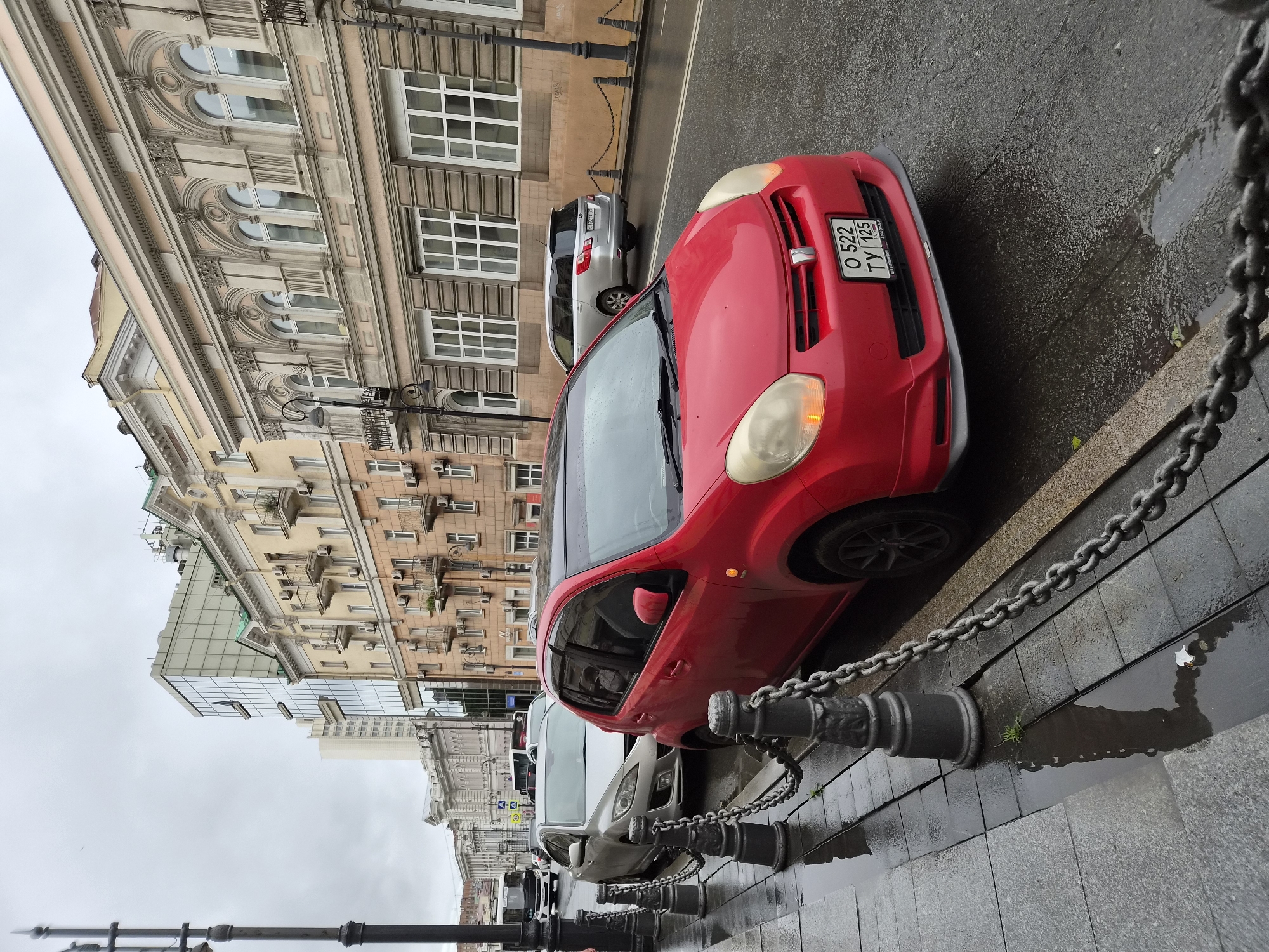 The image shows a street scene in Vladivostok, Russia, on a cloudy day. The foreground features a red car parked along a sidewalk lined with decorative metal posts and chains. The sidewalk is wet and reflects the surroundings, indicating recent rain. Behind the red car, a silver car is also parked. In the background, there are several buildings, some with ornate architectural details and balconies, while others are more modern with glass facades. The street is paved and appears to be wet. Traffic is visible in the distance, including a bus. The overall atmosphere is one of a typical urban street after a rainfall.