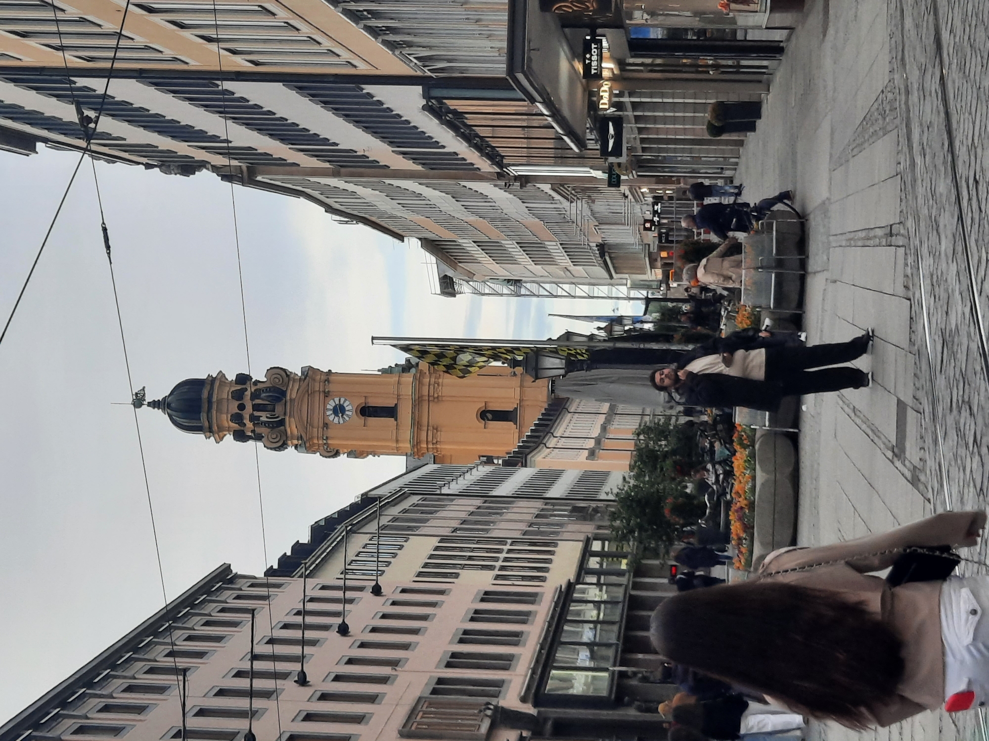 The image depicts a street scene in an urban environment, likely in Europe, characterized by tall, multi-story buildings flanking a pedestrian walkway. A prominent clock tower, with a distinctive orange facade and a dark dome, rises in the background. The street is paved with cobblestones, and tram tracks are visible. Several people are present, walking, standing, and interacting. In the foreground, a woman with long dark hair is seen from behind, wearing a light-colored jacket and white pants. Further down the street, a man in a dark jacket and light-colored shirt is standing. The buildings on either side feature numerous windows and architectural details. The sky is overcast. Businesses with signage are visible along the street, including "TISSOT" and "DoDo". Flower planters with orange and red flowers add a touch of color to the scene. The overall atmosphere suggests a daytime setting with moderate pedestrian activity.