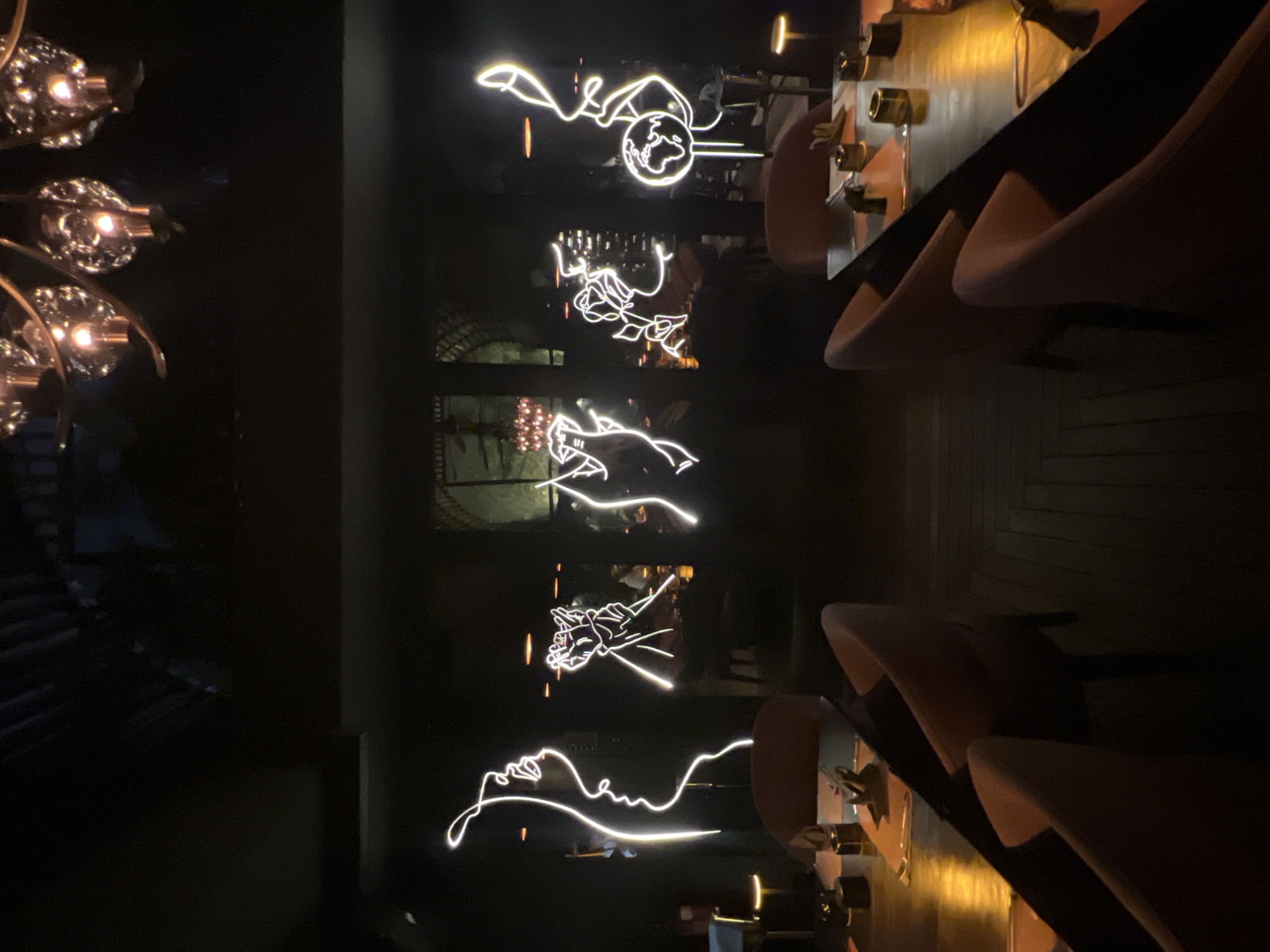 A bar or restaurant in North Kuta, Indonesia is depicted. The image shows a dimly lit interior with modern decor. On the left side of the image, a chandelier with multiple light fixtures hangs down, partially obscuring some of the view. On the wall are stylized neon art, some possibly representing faces or figures in a modern artistic style. Tables and chairs are situated, suggesting a setting for dining or drinks. The chairs are soft, modern, and appear to be comfortable. The lighting creates a contrast between the illuminated neon art and the darker background. The overall scene gives an atmosphere of a chic, contemporary setting.