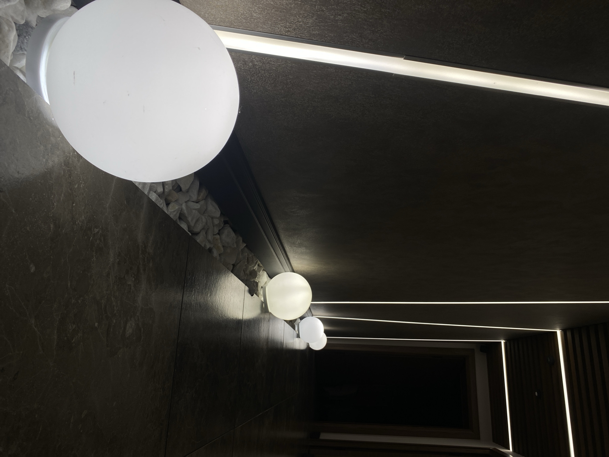 The photograph shows an interior space with a contemporary design featuring a dark color palette contrasted by linear and spherical white light sources. The left side of the frame features a dark, possibly marble or granite, wall with small white stones at the top edge, where the wall meets the ceiling. Attached to the wall are three spherical white light fixtures, varying in size and diminishing in perspective towards the back of the space. The ceiling is dark and incorporates linear recessed lighting elements. In the distance, there is a dark window or opening framed by light wood. To the right of the frame are dark wooden slats which contribute to the ambient lighting. There is no information that could be used to determine if the image was taken in Votkinsk, Russia. There are no people present in the image.