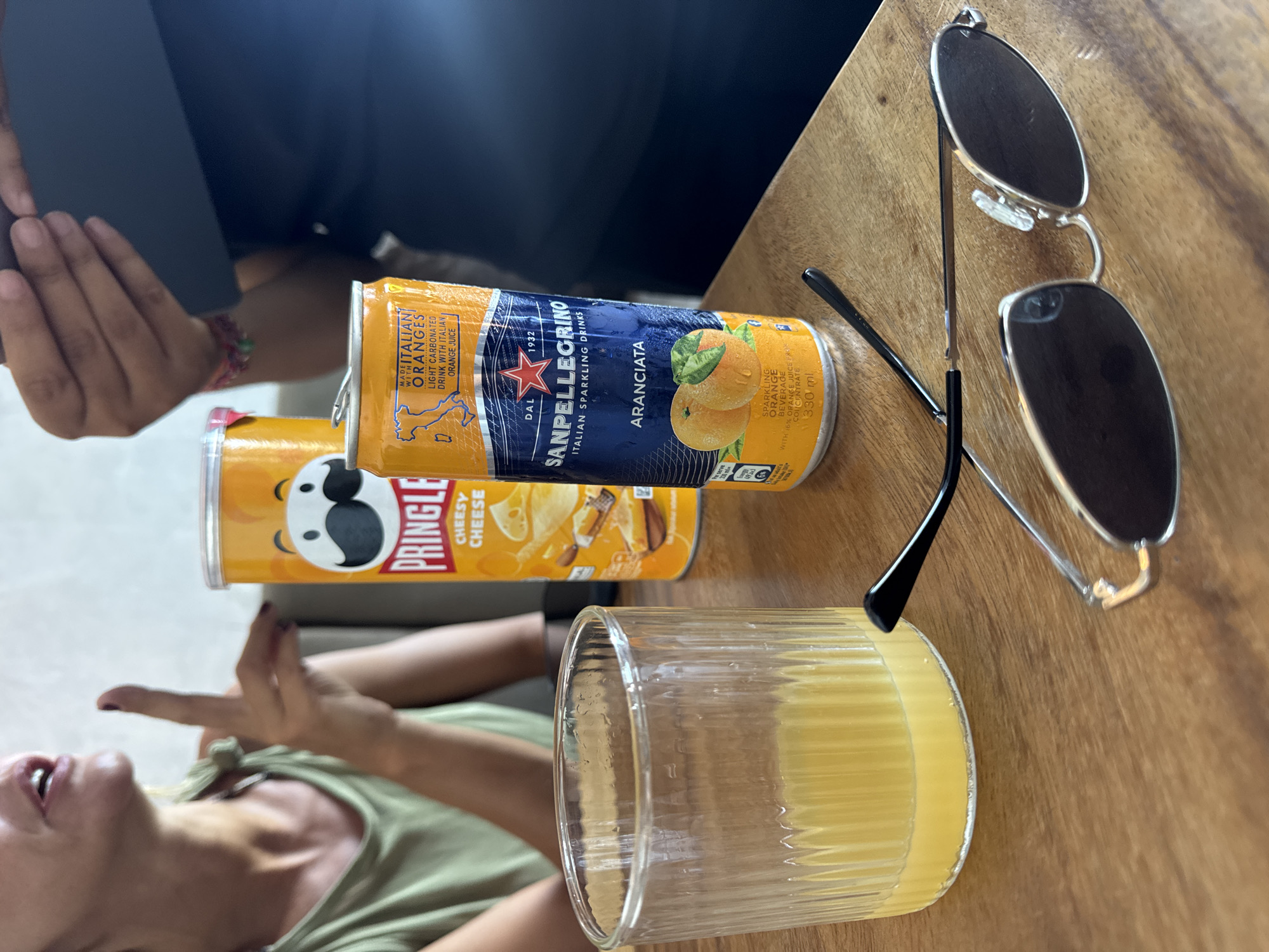 A table setting features a can of Pringles Cheesy Cheese, a can of San Pellegrino Aranciata (Italian Sparkling Orange Drink), a ribbed glass filled with a yellow liquid, and a pair of sunglasses. Two people are partially visible in the background. The setting is in Kuta Selatan, Indonesia.