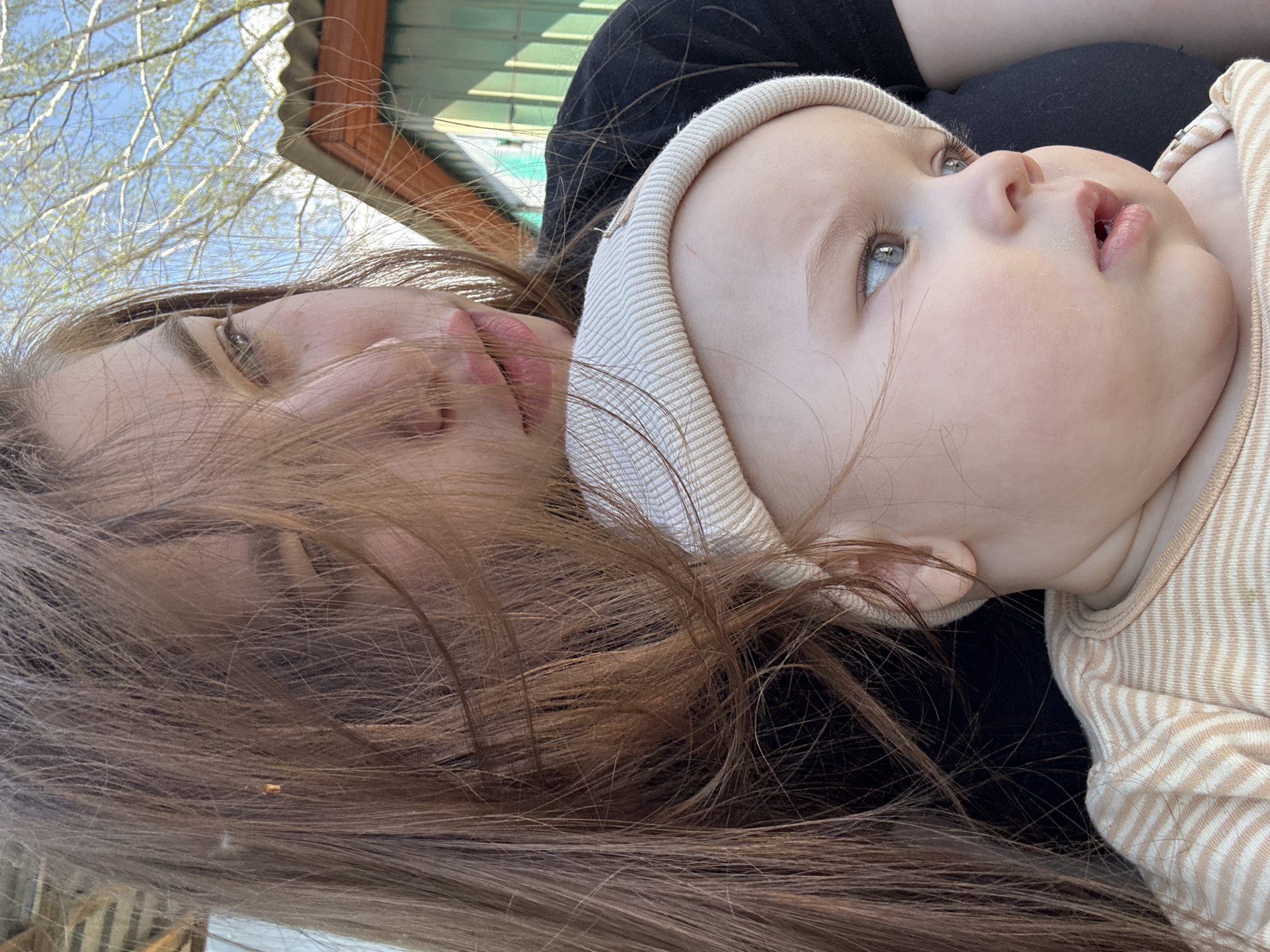 A medium shot of a woman lying on her side outdoors with a baby on her chest. The woman has long, brown hair that falls across her face. Her eyes are closed, and she is looking at the baby. The baby is wearing a light-colored cap and a striped onesie. The baby's eyes are open, and it is looking up and to the right. The background is blurred and shows a green building, blue sky, and tree branches. The location is potentially Мещёры, Russia.