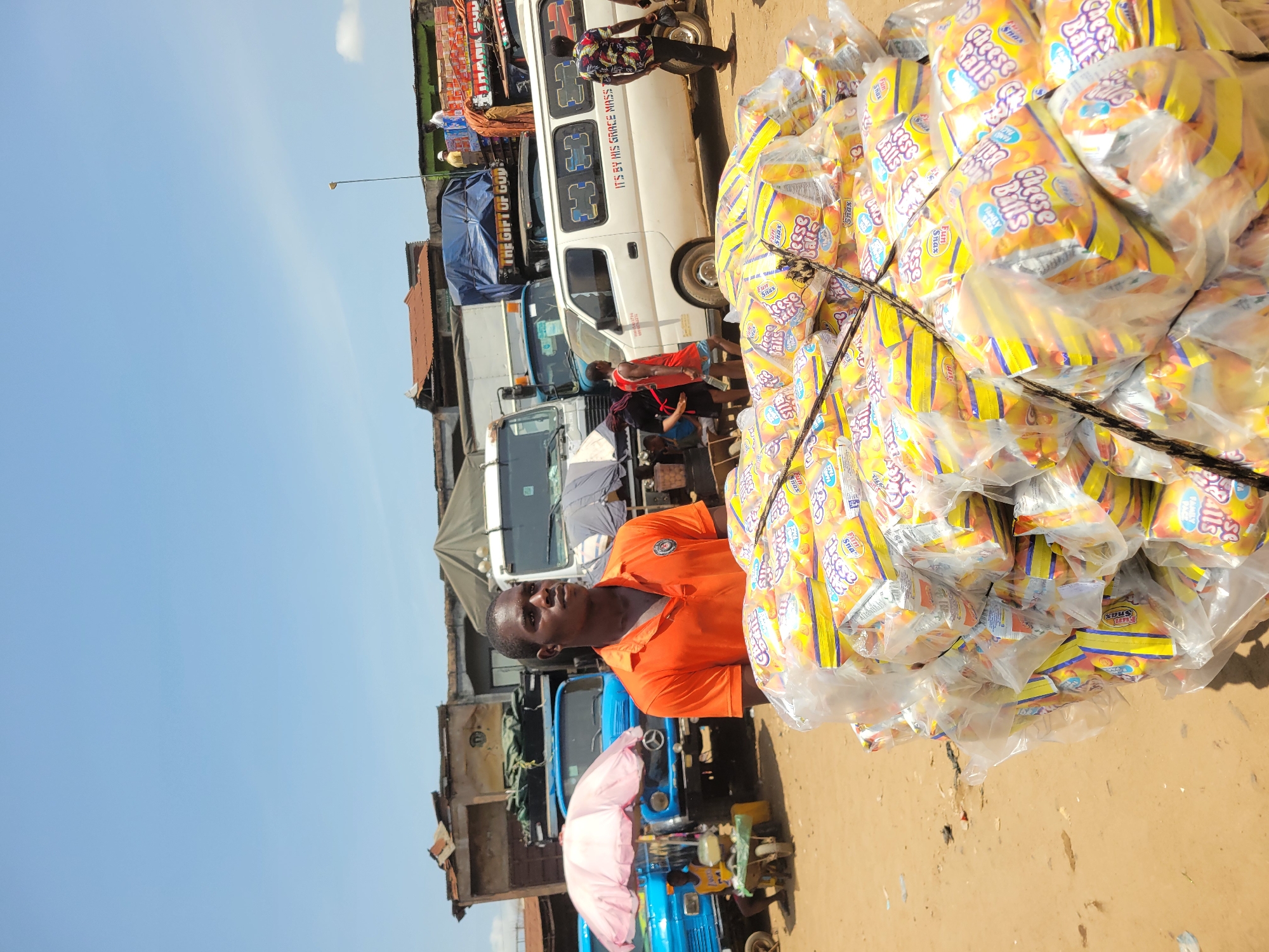 The image captures a bustling outdoor market or commercial area, likely in Onitsha, Nigeria, under a bright, clear blue sky on a sunny day. In the immediate foreground, a man wearing an orange polo shirt stands, looking directly at the camera with a neutral expression. To his right, a large, tall stack of clear plastic bags filled with smaller, yellow and orange packages of snacks dominates the right side of the frame. The packages are visibly labeled "Cheese Balls" and "Fun Snax," some also showing "FAMILY" or "Star" branding.
The background reveals a lively scene of transportation and commerce. Several vehicles are parked or moving, including a white passenger van with "IT'S BY HIS GRACE MAS" (likely "MASTER") and "THE GIFT OF GOD" text visible on its side. Various trucks, some covered with tarpaulins, are also present, suggesting the movement of goods. A blue three-wheeled utility vehicle, possibly a Keke NAPEP, with a pink umbrella attached, is positioned further back near the man in orange. Scattered people can be seen in the mid-ground, engaging in daily activities typical of a busy market, such as walking, standing, and interacting near the vehicles. Buildings with simple structures and corrugated metal roofs line the horizon, completing the impression of an active urban or semi-urban commercial hub. The ground is unpaved and appears dusty, indicating an outdoor, functional environment.