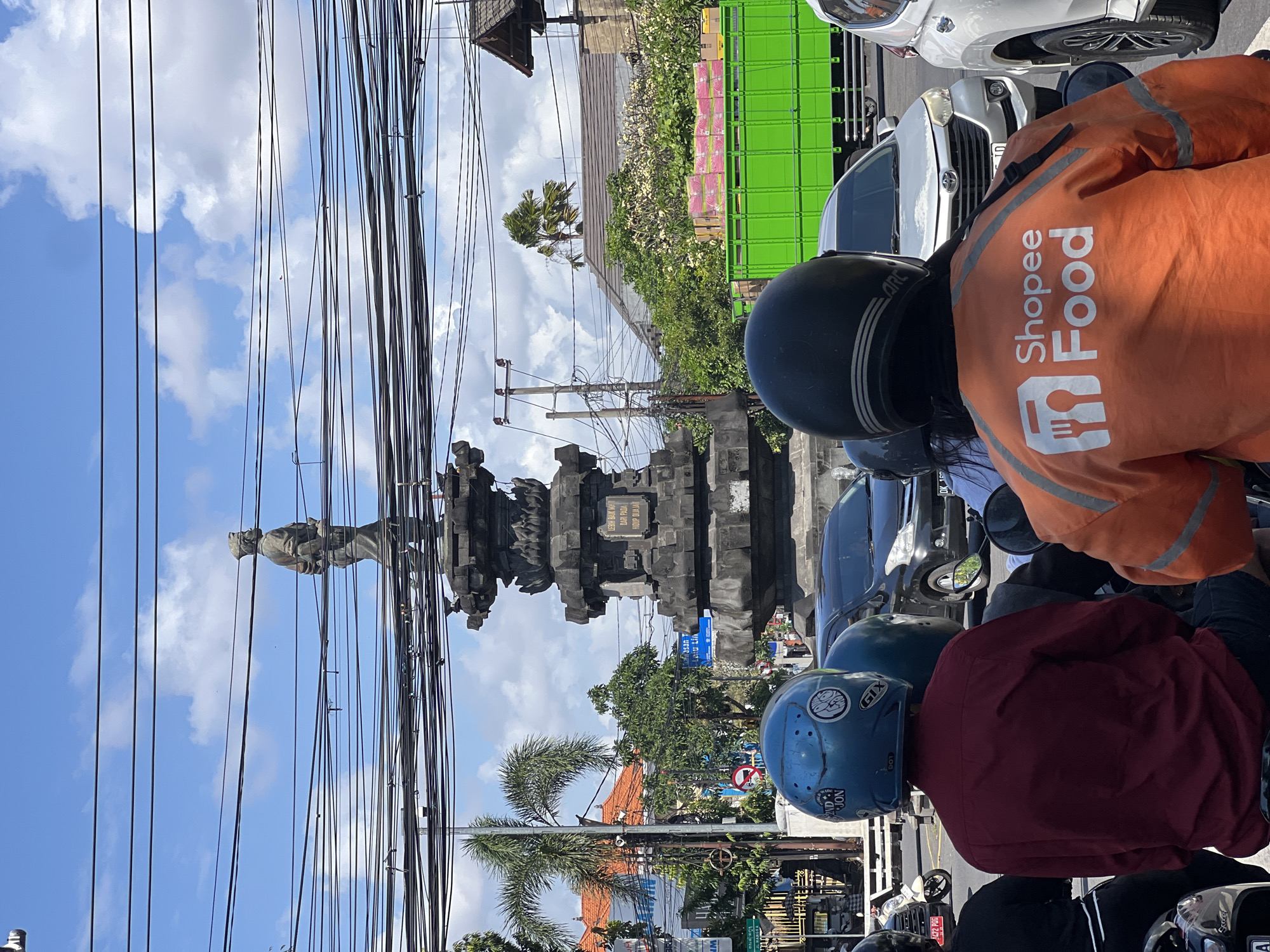 The image captures a street scene in Denpasar, Bali, Indonesia, on a sunny day with scattered clouds. A prominent stone monument, likely a historical marker or a fountain, stands in the center of the frame, adorned with intricate carvings and topped with a statue of a man in uniform. Overhead, a dense network of electrical wires crisscrosses the sky, obscuring parts of the blue sky and the monument.
In the foreground, three individuals are seated on motorcycles, their backs to the camera. The person on the left wears a red long-sleeved shirt and a blue helmet adorned with stickers. The central figure is wearing an orange jacket with the "Shopee Food" logo prominently displayed on the back, indicating they are a food delivery courier. The person on the right, partially visible, also wears a helmet.
The background reveals a bustling urban environment. To the left of the monument, palm trees and buildings with terracotta roofs are visible. To the right, a large green truck is parked, carrying boxes. Cars are interspersed in the scene, suggesting traffic on the road. The overall impression is of a typical street intersection in a tropical city, characterized by a blend of cultural landmarks, modern infrastructure, and daily life.