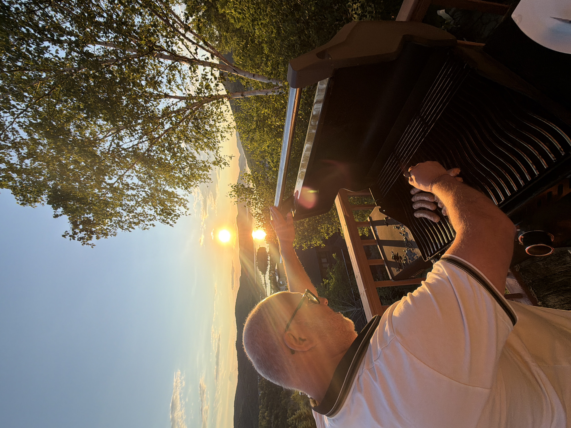 A man with glasses and a light-colored polo shirt is grilling sausages on a barbecue on a wooden deck during sunset. The sun is setting over a body of water, casting a warm glow on the landscape of hills and trees. A birch tree with green leaves is visible to the right of the frame. The setting appears to be a residential area in the city of Mont-Tremblant, Canada, given the natural mountainous terrain and the presence of a lake or large river. The man is leaning over the grill, his right hand reaching to adjust a sausage. The grill is open, and several sausages are visible on the grates. The sky is clear with a few scattered clouds.