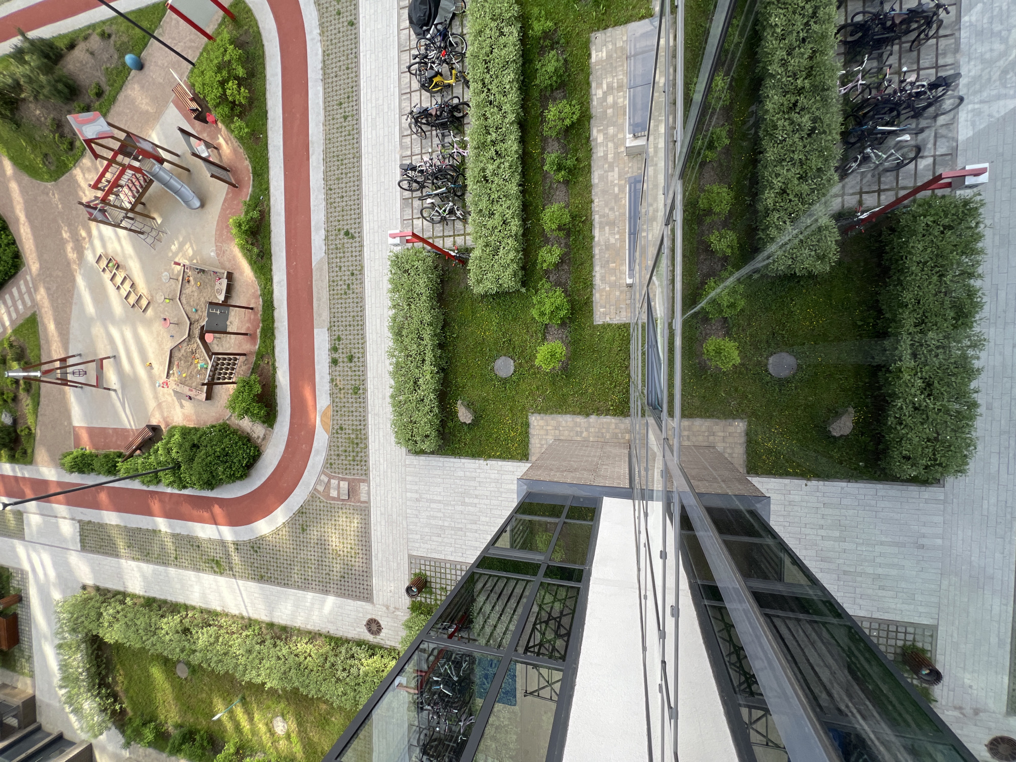An aerial, downward-looking shot captured from a high-rise building showcases a modern urban courtyard. The image is tilted, offering a unique perspective of the ground below.
The scene features a well-maintained outdoor space with distinct zones. On the left, a children's playground is visible, equipped with red and grey climbing structures, a slide, sandboxes, and various small wooden steps and platforms. Paved pathways and a prominent red running track curve through this area.
Adjacent to the playground, a strip of manicured green lawn is bordered by dense hedges. Several bicycles are neatly parked in multiple racks along a paved walkway, indicating a dedicated cycling area. Further to the right, more green spaces with small bushes and grass are interspersed with paved sections.
A prominent feature is the reflective glass facade of the building from which the photo is taken, occupying the right side of the frame. This facade mirrors the sky and parts of the adjacent ground, creating abstract reflections. Below this reflective surface, a glass-covered walkway or entrance structure is also visible. The ground cover includes various textures: light-colored pavers, grey brickwork, and grass with visible manhole covers.
No people are visible in the frame. The lighting suggests it is daytime, with clear skies. The overall impression is one of a clean, organized, and modern residential environment in Saint Petersburg, Russia, designed for recreation and community living. No visible text is present in the image.