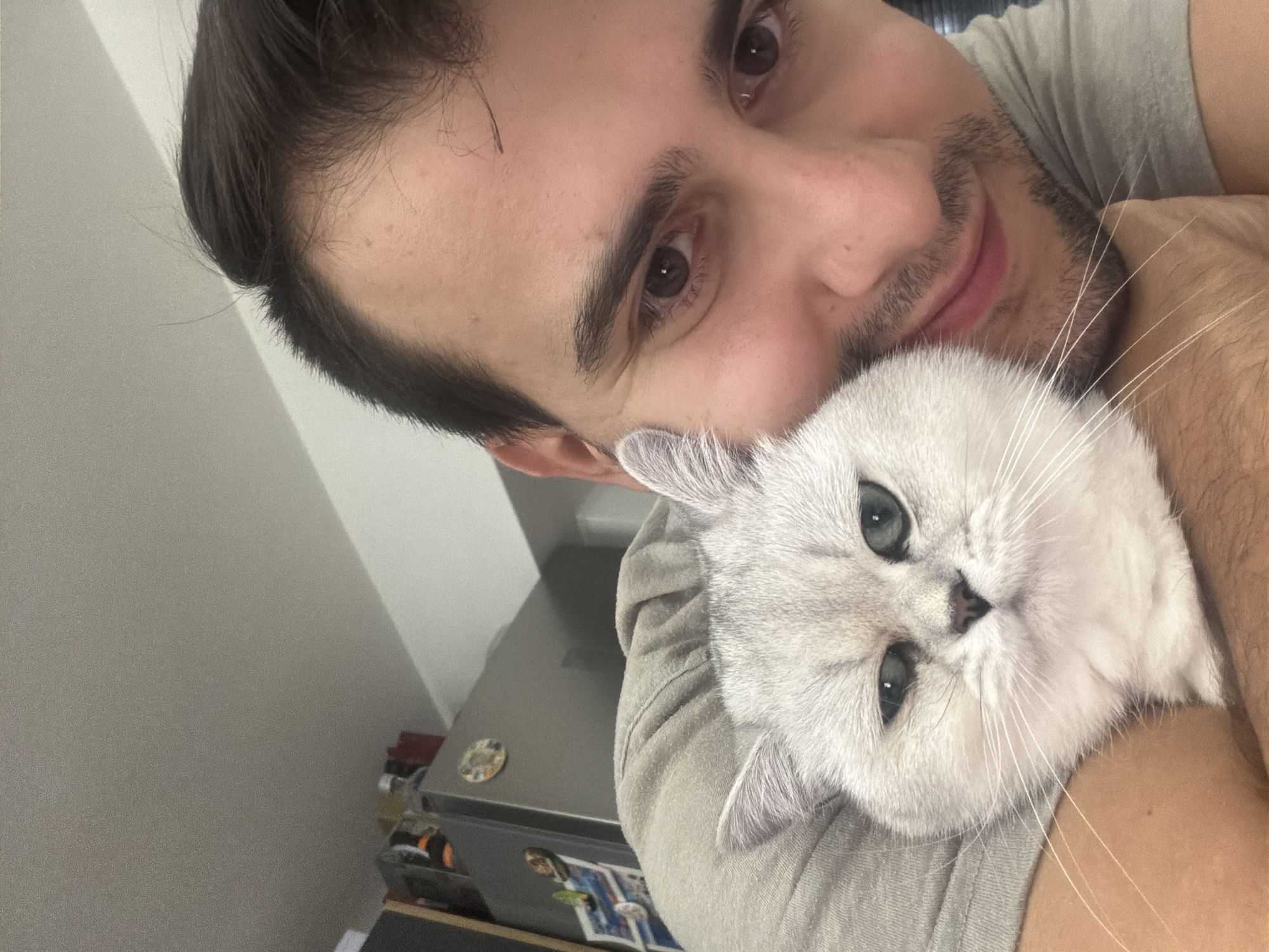 A man is holding a white cat close to his face. The man has dark hair and brown eyes, and he is smiling slightly. The cat has blue eyes and white fur. The man is wearing a gray t-shirt. In the background, there is a gray surface, possibly a refrigerator, with magnets. The photo was not taken in Burgas, Bulgaria.