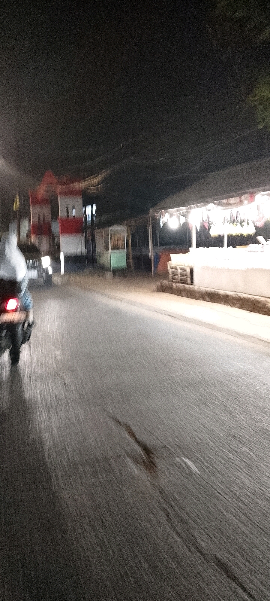 The image depicts a night-time scene on a street in Depok, Indonesia. The perspective is from a moving vehicle, likely a motorcycle, given the presence of a motorcycle's taillight in the lower left corner. The road surface is wet and reflects the ambient light, showing streaks from the motion blur. On the right side of the street, there are several stalls or shops, one of which is illuminated by bright lights and appears to be selling items displayed on a counter. These stalls are covered by awnings. In the background, to the left, a building with red and white stripes is visible. A person wearing a white hooded jacket is on the motorcycle in the foreground. The overall impression is that of a busy street scene at night with commercial activity.