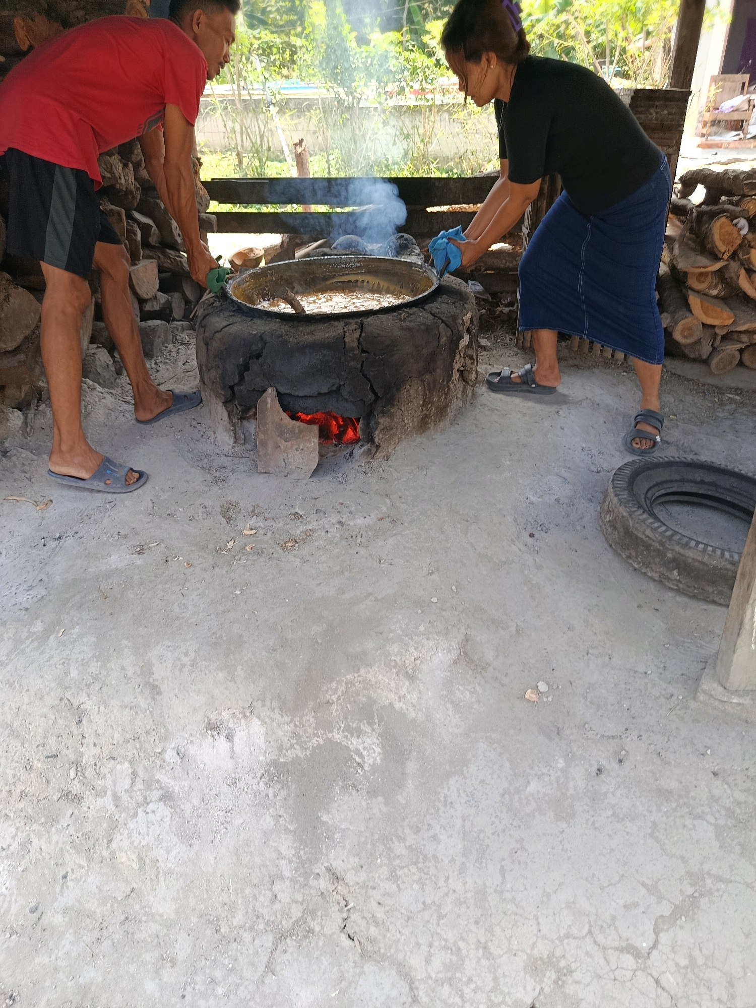 In Sarwadadi, Indonesia, two individuals are engaged in cooking over an open fire. A man in a red t-shirt and black shorts stands to the left, leaning over a large cauldron placed within a makeshift oven constructed from a tire. He is holding a green tool, possibly for stirring the contents of the cauldron. To the right, a woman wearing a black t-shirt and a blue skirt also participates, holding a blue cloth. The fire beneath the cauldron is visible, emitting smoke. The ground is a mixture of concrete and dirt, with scattered debris. In the background, a stack of firewood and lush greenery are present, suggesting an outdoor or semi-outdoor setting. A discarded tire lies on the ground near the woman.