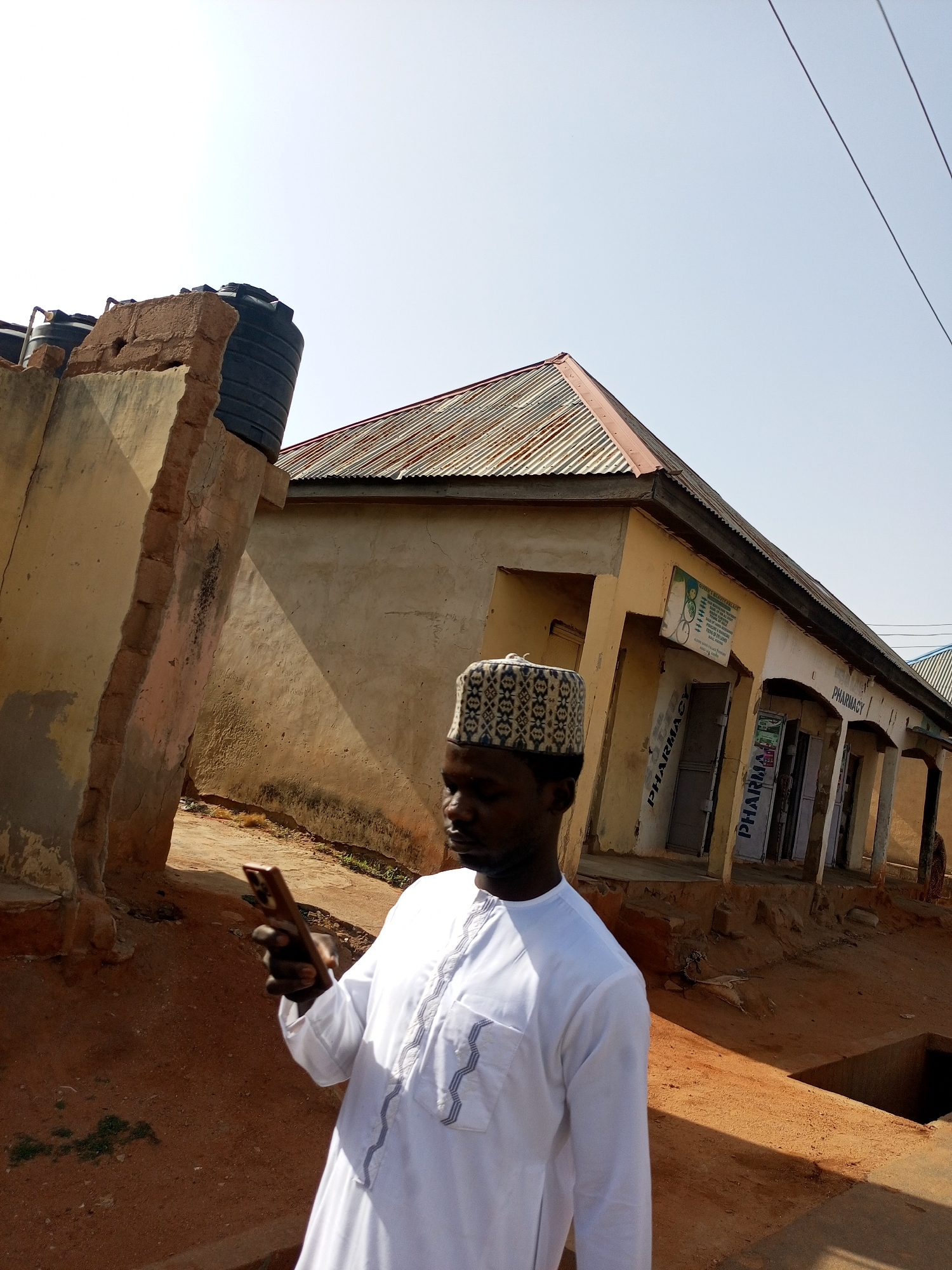 A man in a white robe and patterned cap holds a smartphone in his left hand, looking down at the screen. He stands on reddish-brown dirt in front of a single-story, tan building with multiple entrances labeled "PHARMACY." The building has a corrugated metal roof and a small sign is visible above one of the doors. To the left, a crumbling concrete structure with a black water tank on top is observed. Overhead power lines cross the sky. The location is identified as Tempure, Nigeria.
