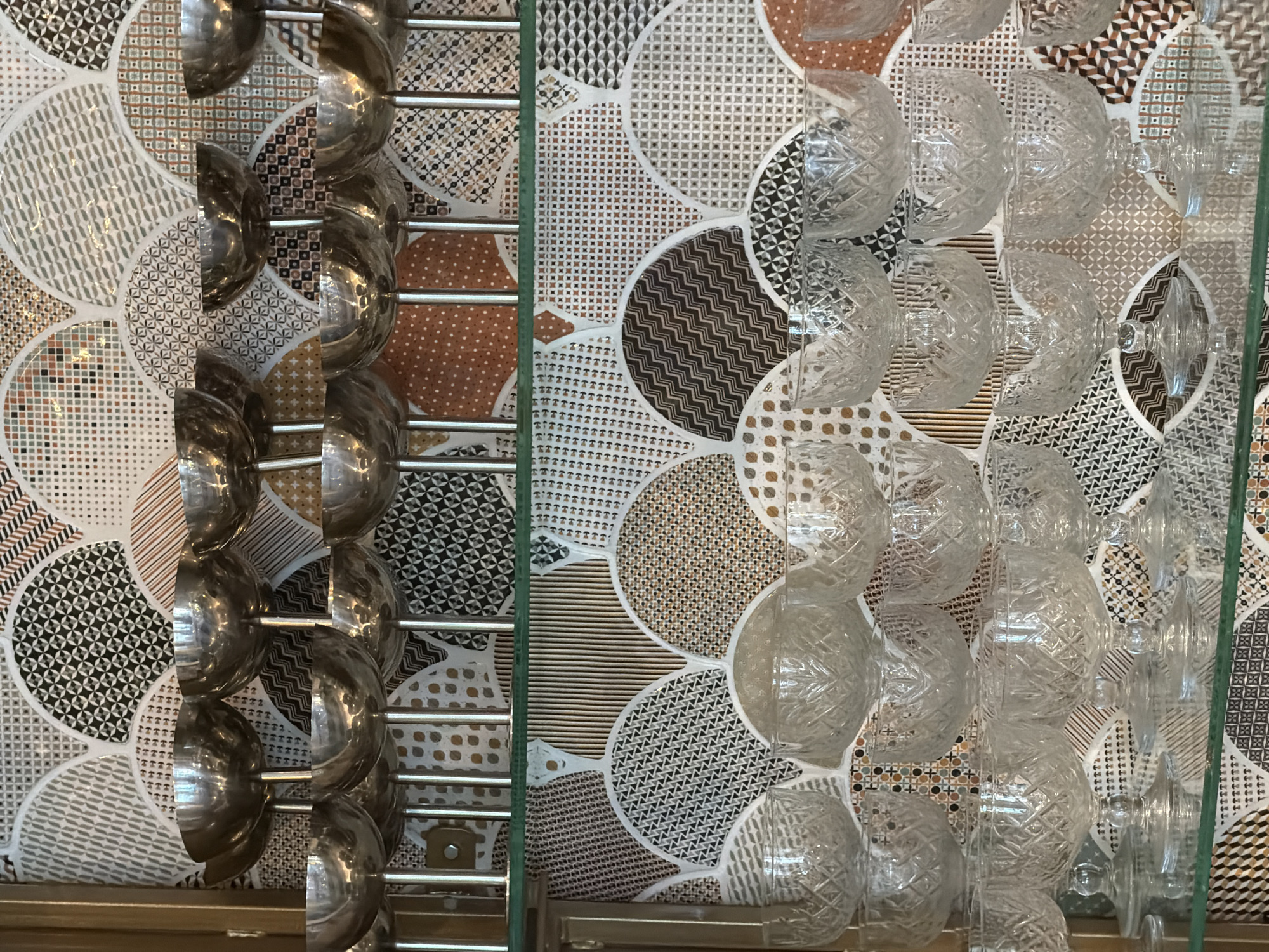 The image depicts a close-up of a kitchen utensil rack in Radovići, Montenegro. On the left, stainless steel ladles hang from a metal rack. To the right of a glass divider, clear glass bowls and what appears to be shot glasses are suspended from another rack. The backdrop behind both racks consists of a decorative wall tile pattern featuring a repeated circular design with varying geometric fills and a color palette of browns, whites, and grays. The bottom of the image shows a wooden or laminate surface.