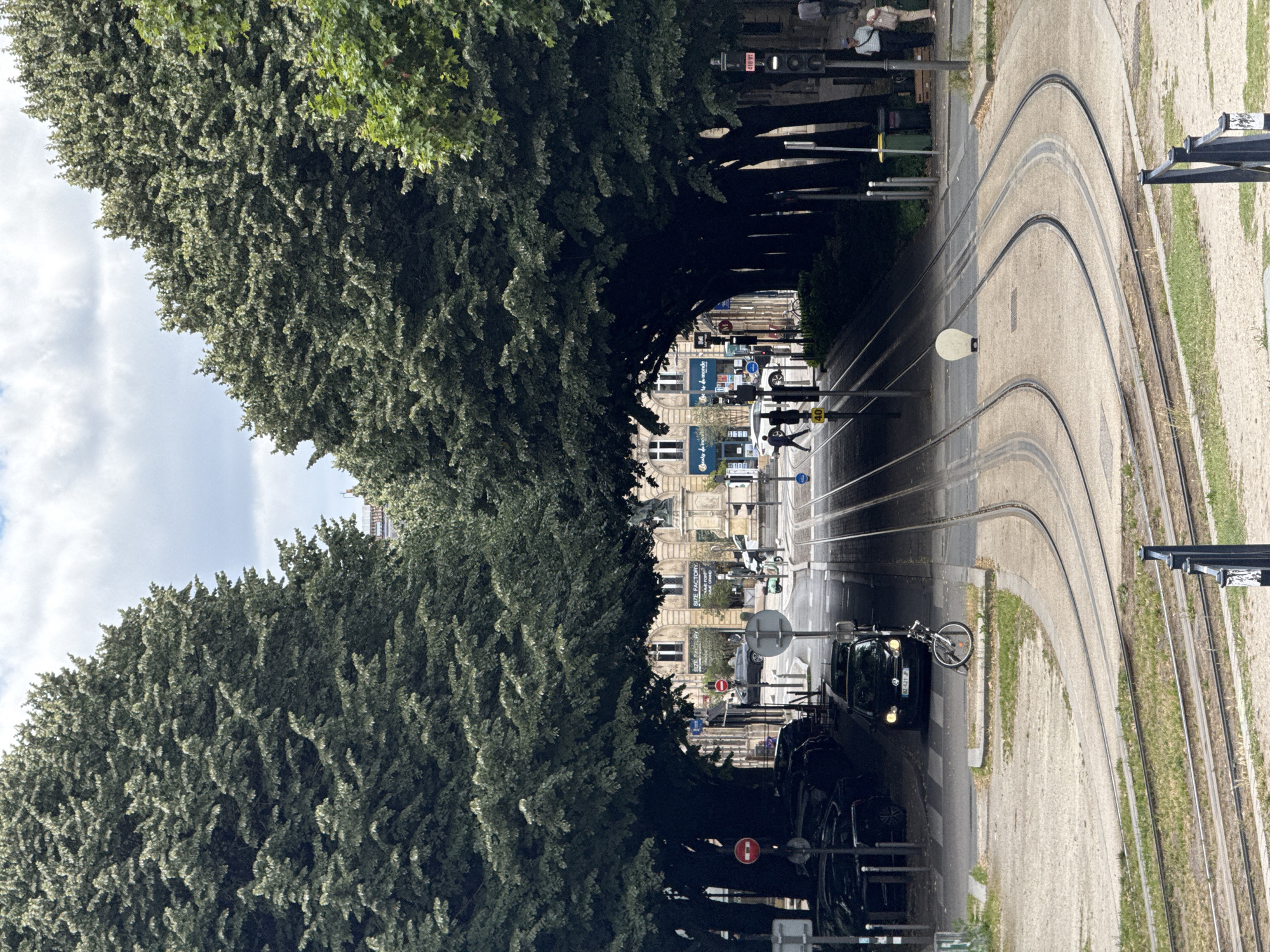 The image captures a street scene in Bordeaux, France, likely in a park or along a major thoroughfare lined with mature trees that create a canopy effect. Tram tracks are prominently featured, curving through the foreground and disappearing into the distance. A black car is parked on the left, and a bicycle is leaning against a low barrier. Beyond the parked car, tram tracks continue and merge with a street. Several people are visible on the street, including two individuals crossing a road and two people standing on a sidewalk near a traffic light. Buildings with classical architecture line the street in the background. The sky is partly cloudy, suggesting overcast weather. The overall impression is one of an urban environment with significant green spaces and public transportation infrastructure.