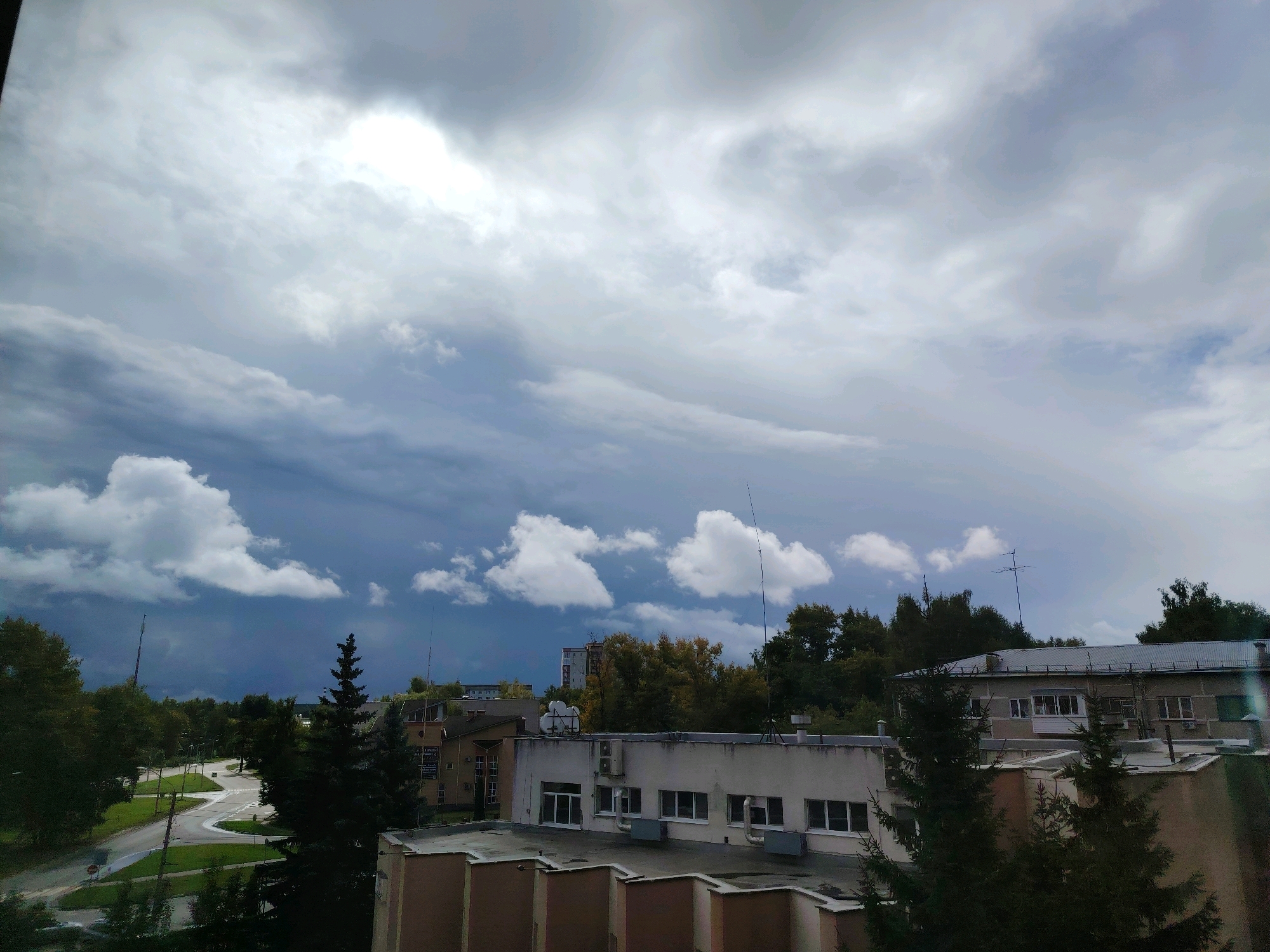 The image captures a view from an elevated vantage point in Konakovo, Russia, looking towards a partially cloudy sky and a residential area. The sky is dominated by large, dramatic clouds, with patches of brighter light suggesting the sun is attempting to break through. In the middle ground, a road network is visible, with cars driving on it. Several buildings are present, including a light-colored building with multiple windows and air conditioning units on its facade, and other structures with darker roofs and walls. Trees, some appearing as evergreens and others with autumnal foliage, are scattered throughout the scene, bordering the road and surrounding the buildings. The overall impression is of an overcast day in a suburban or semi-urban environment.