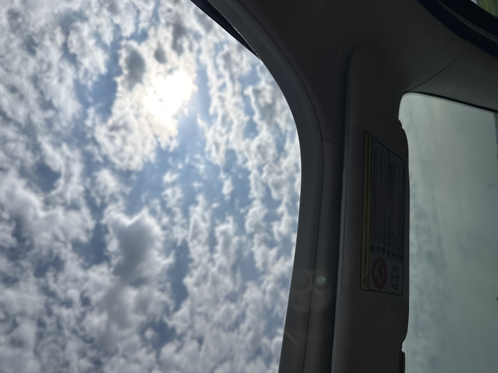 The image captures a view through a car's sunroof on a cloudy day. The sun is visible through breaks in the scattered clouds, casting a bright glow. The sunroof itself is visible in the foreground, with its interior trim and a warning label attached to the visor. The warning label contains text in Italian and several pictograms, including one depicting a person with a seatbelt and another showing an open book. The overall impression is one of travel, with the vast sky and clouds suggesting an open road. There are no people or specific landmarks visible that would allow for identification of the city of Longueil, France.