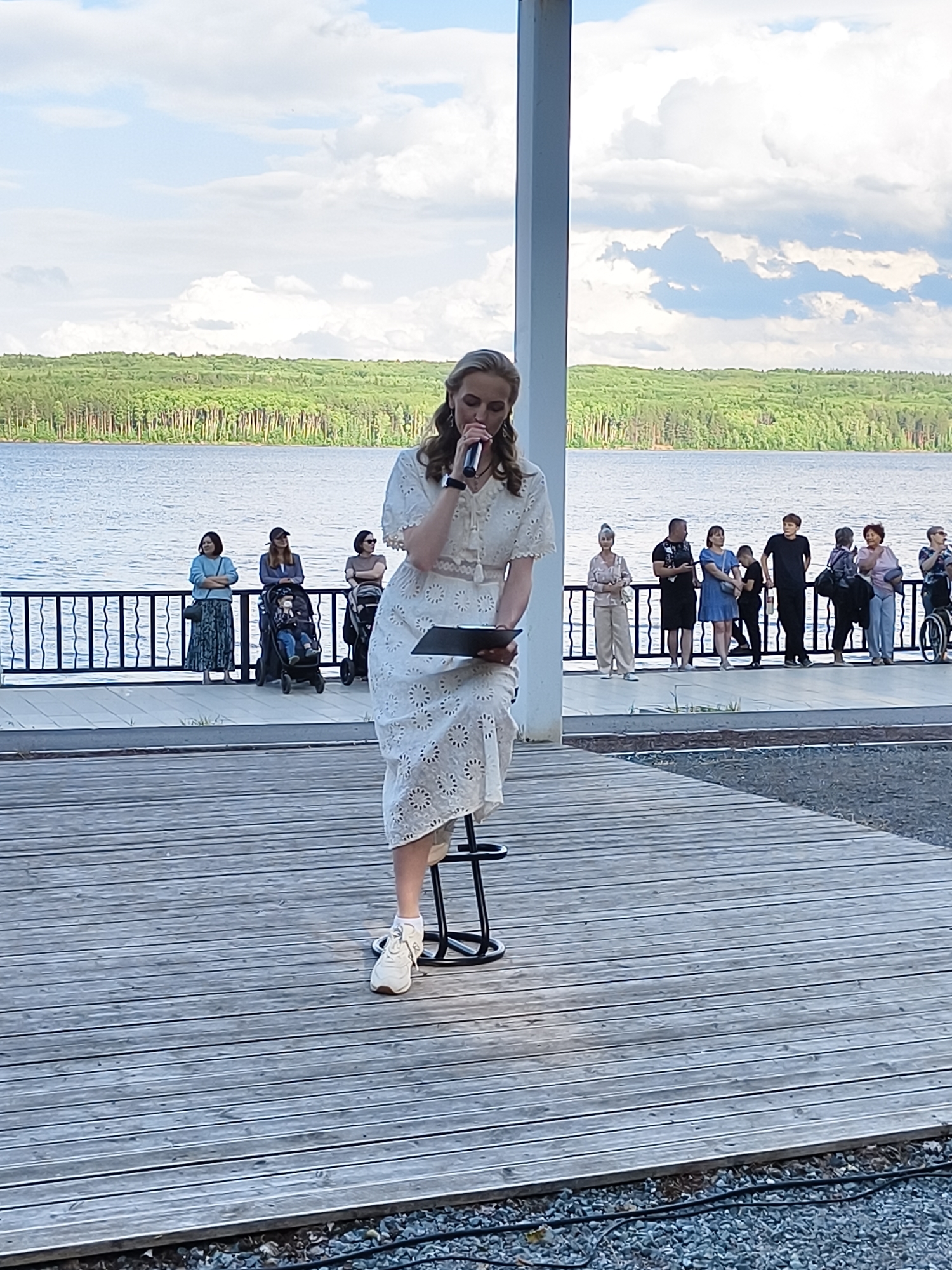 The image captures an outdoor event, likely near a body of water, possibly on the Kama River embankment in Chaikovsky, Russia. In the foreground, a woman in a white lace dress and sneakers stands at a microphone, holding a tablet, positioned on a stool. She appears to be addressing an audience. Behind her, a railing separates the wooden deck from a paved area where a group of people are gathered, some standing and observing, others with strollers. The background features a calm body of water with a tree-lined shore stretching across the horizon under a cloudy sky.