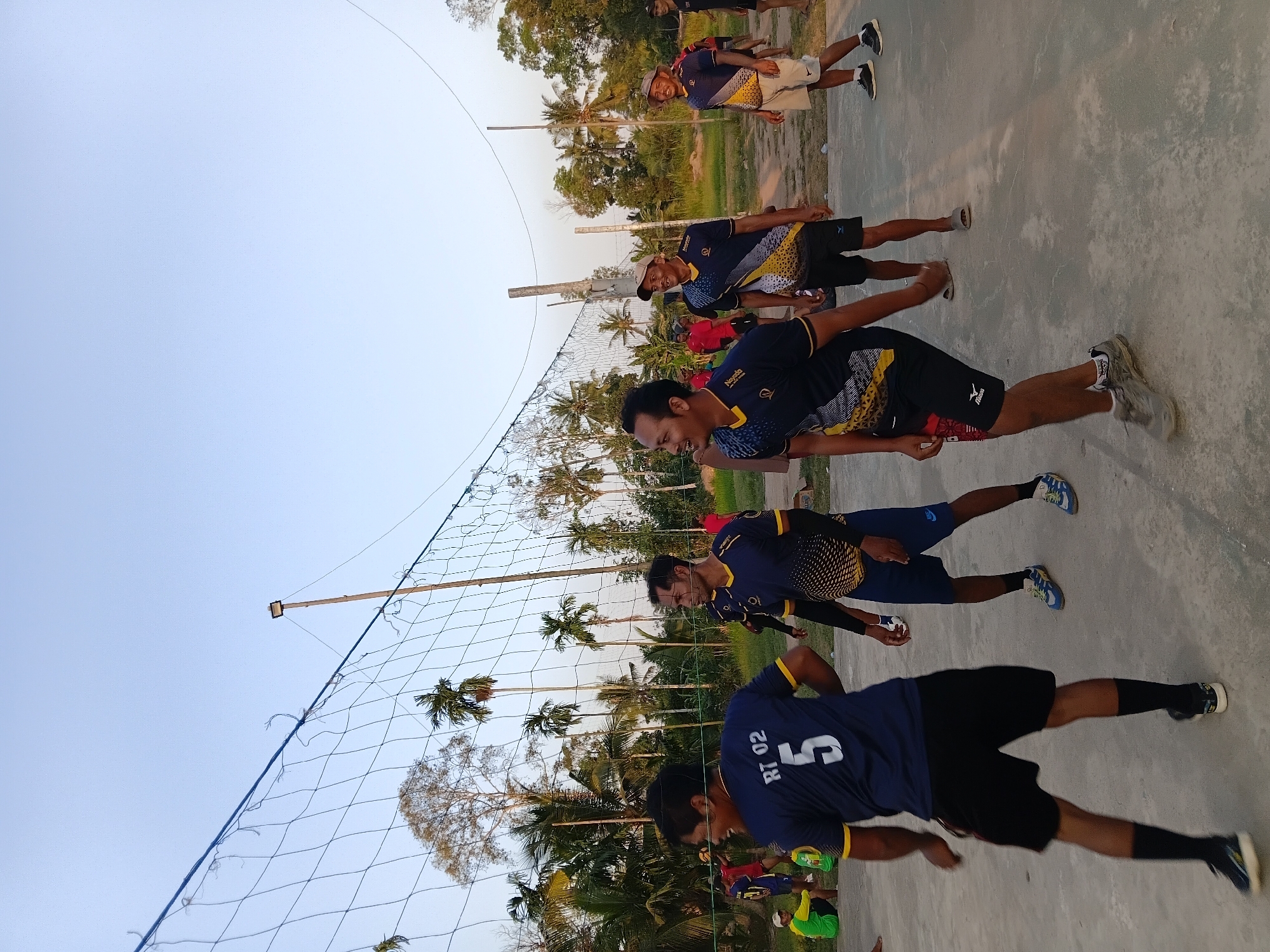 In Margo Rukun, Indonesia, a group of men are playing volleyball on a dusty court outdoors. A net is stretched across the court, with two prominent poles supporting it. Several players are visible in action, wearing athletic shorts and jerseys. One player in the foreground wears a dark blue jersey with yellow accents and the number "5" prominently displayed on the back, along with "RT 02." Another player nearby has a similar jersey with a black and yellow patterned design. Other individuals are scattered around the court, some observing and others participating. The background features a line of palm trees and other vegetation under a clear sky, suggesting a tropical or subtropical environment. A single floodlight is mounted on a tall pole behind the net. The overall scene depicts a casual game of volleyball in a rural or semi-rural setting.