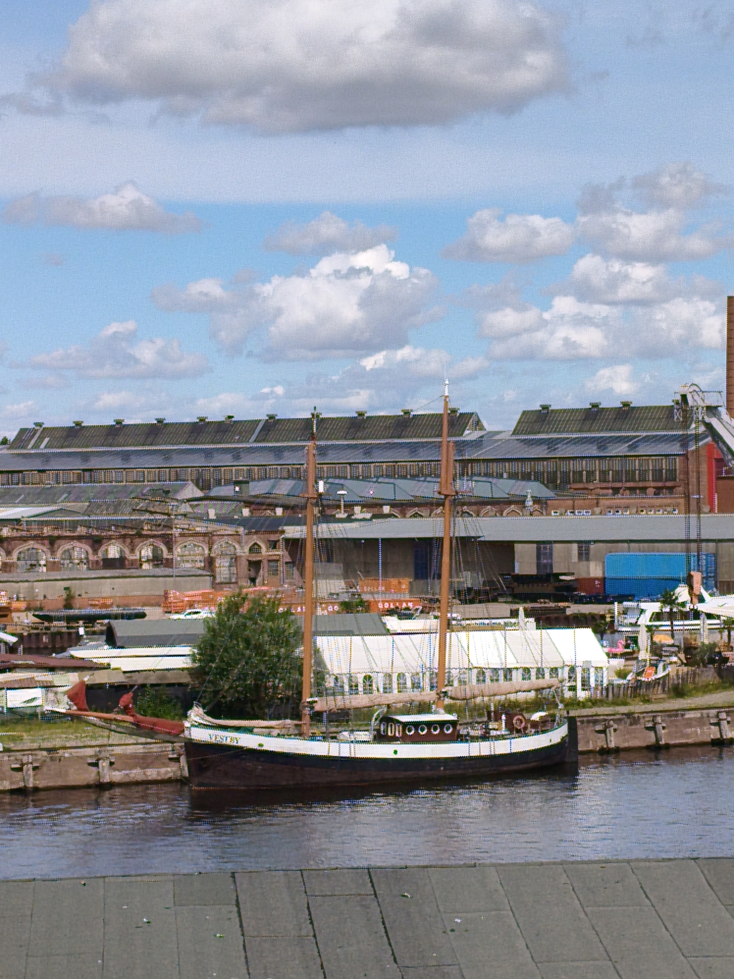 The image depicts a boat named "VESTBY" docked in a harbor with industrial buildings in the background, likely in the city of Lübeck, Germany, given the characteristic architecture of the warehouses. The boat, a two-masted schooner with a dark hull and white superstructure, is moored alongside a concrete quay. In the background, large, multi-story buildings with arched windows and long, sloped roofs dominate the scene, suggesting a historical industrial or maritime area. A large blue shipping container is visible near the quay. The sky is partly cloudy with white cumulus clouds against a light blue expanse. The foreground shows a grey paved walkway, possibly part of the harbor infrastructure. No people are clearly visible in the image, and the overall atmosphere is one of stillness and a working port environment.