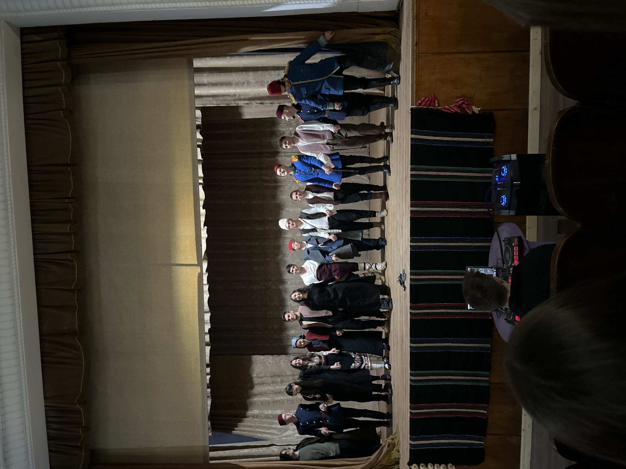 A stage performance is captured in a photograph taken in Voiniagovo, Bulgaria. The stage has a brown curtain backdrop and floor coverings with colorful stripes. A group of approximately fifteen people are standing in a line on the stage, dressed in what appears to be period or traditional costumes. The costumes include red fez hats, military-style jackets, dresses, and vests. An audience is partially visible to the right of the stage, with some viewers holding phones. A sound system or speaker is also visible in the background.