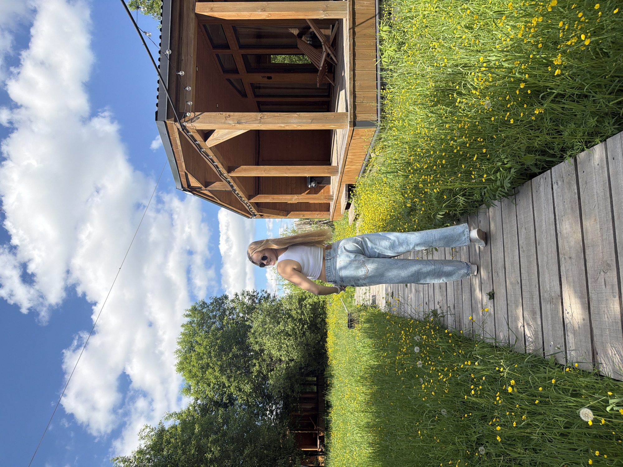 A young woman is captured posing on a wooden walkway in a bright, natural outdoor setting, likely in Vozhoiskii, Russia. She stands mostly sideways to the camera, with her head turned to look directly at it, displaying a slight smile. She has long, straight light-colored hair and is wearing a white sleeveless crop top, light blue baggy jeans, and sunglasses.
The wooden walkway stretches into the distance, bordered by a dense field of vibrant green grass profusely covered with small yellow flowers, possibly buttercups or similar wildflowers. Beyond this floral expanse, a line of large green trees with full canopies forms the background on the left side of the image.
To the woman's left, a rustic wooden building, resembling a cabin or gazebo, stands with a dark roof. It features a covered porch-like area with wooden posts and visible window panes. String lights with clear, round bulbs are strung along the eaves of the building, adding a decorative touch. A wooden chair is discernible inside the structure.
The scene is bathed in bright daylight under a clear blue sky dotted with scattered white, puffy clouds. The overall mood is pleasant and idyllic, suggesting a serene and relaxed environment. No visible text is present in the image.