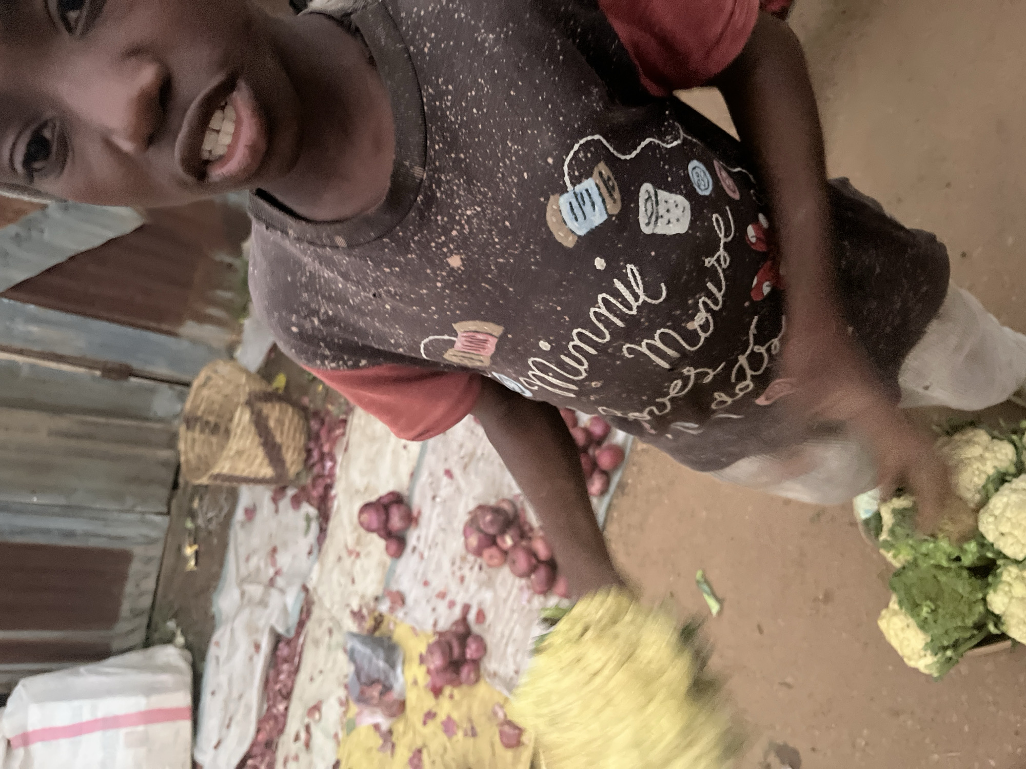 A young individual is standing in a market setting, likely in Gura Top, Nigeria. The individual is wearing a dark t-shirt with a red collar and sleeves. The t-shirt has a graphic design that includes the words "Minnie Mouse" in white script, along with cartoonish illustrations of sewing-related items like spools of thread and thimbles. The individual's mouth is slightly open, revealing their teeth. In front of the individual, there is a collection of cauliflowers in a woven basket. To the left and in the background, there are piles of red onions scattered on a yellow tarp and a white cloth. A large white bag with pink stripes is visible in the far left background, and a woven basket is situated behind the onions. The ground appears to be dirt or sand. The overall scene depicts a market stall with produce for sale.