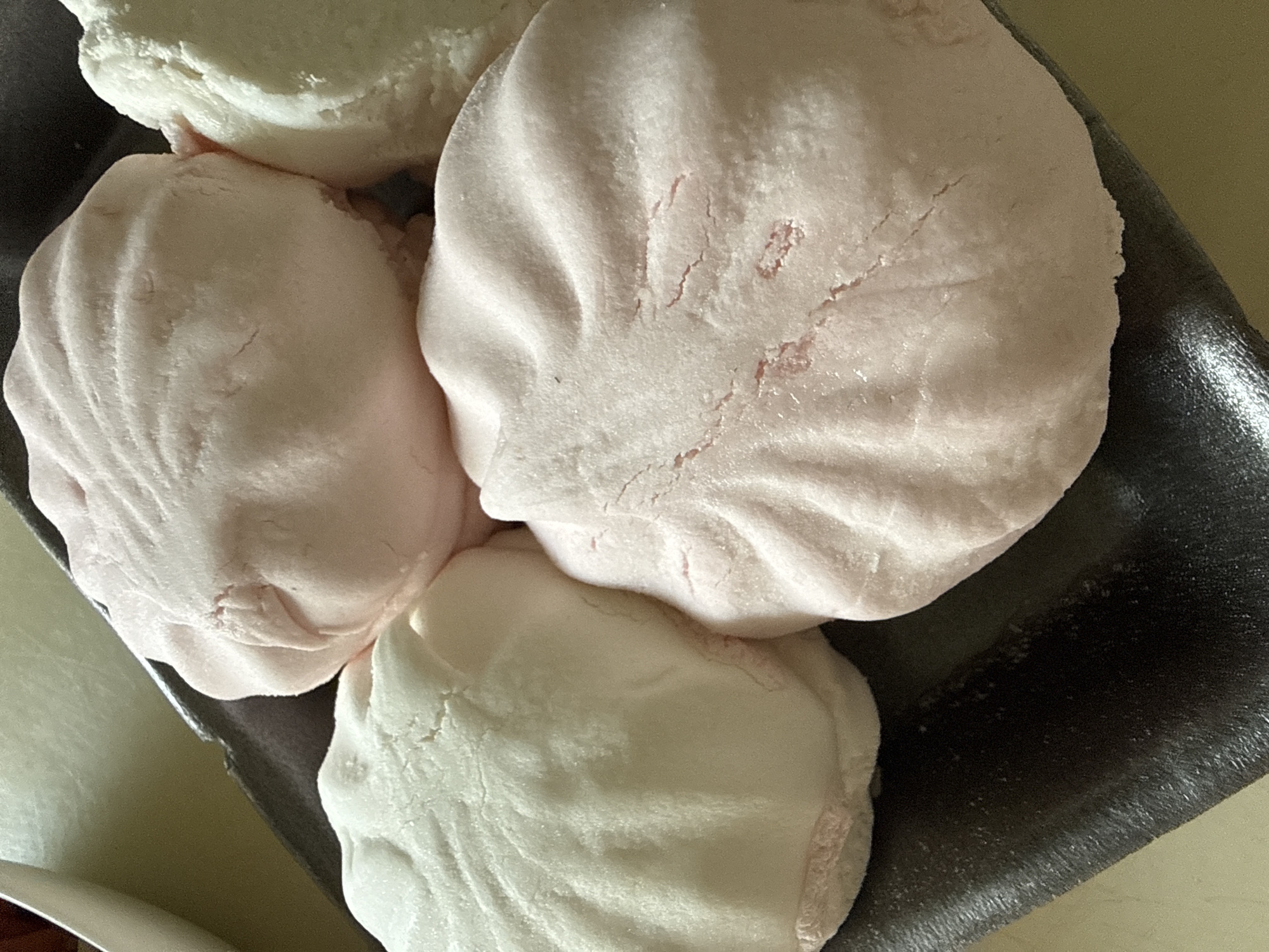 This is a close-up, high-angle, eye-level shot of four large, pastel-colored, irregularly shaped meringues or large marshmallows resting on a dark, textured plastic tray. Two meringues are pale pink, and two are white. The meringues have a soft, pillowy appearance with visible creases and slight cracks on their surfaces, suggesting a powdered or sugary coating. The lighting is bright, casting soft shadows and highlighting the textures of the meringues. The background consists of a light-colored, flat surface and a hint of a box or container. The scene appears to be indoors, likely in a domestic setting. There are no people visible, and no specific details indicate a location in Okhtyrka, Ukraine.