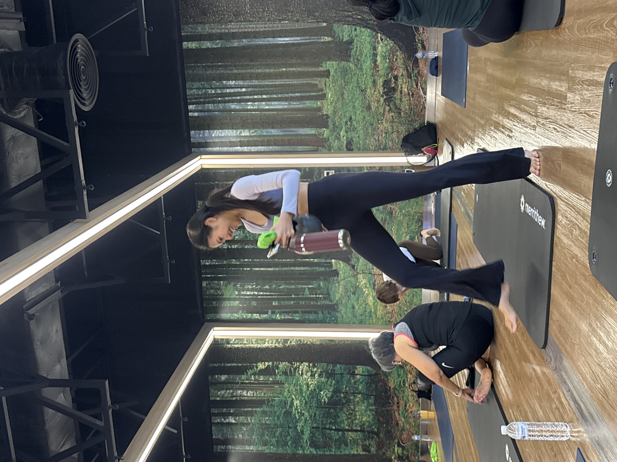 In a yoga studio with a forest mural backdrop, a woman in a white long-sleeved crop top and dark leggings stands on a yoga mat, holding a maroon water bottle and a green object. Her left leg is bent at the knee and her right leg is extended forward. To her left, an older woman with gray hair wears a black tank top and dark leggings, sitting on a mat with her legs crossed. Another person is partially visible in the background, kneeling on a mat. The floor is wooden, and several yoga mats are laid out. The lighting appears to be from overhead fluorescent fixtures. The location is specified as Nong Pa Khrang, Thailand.