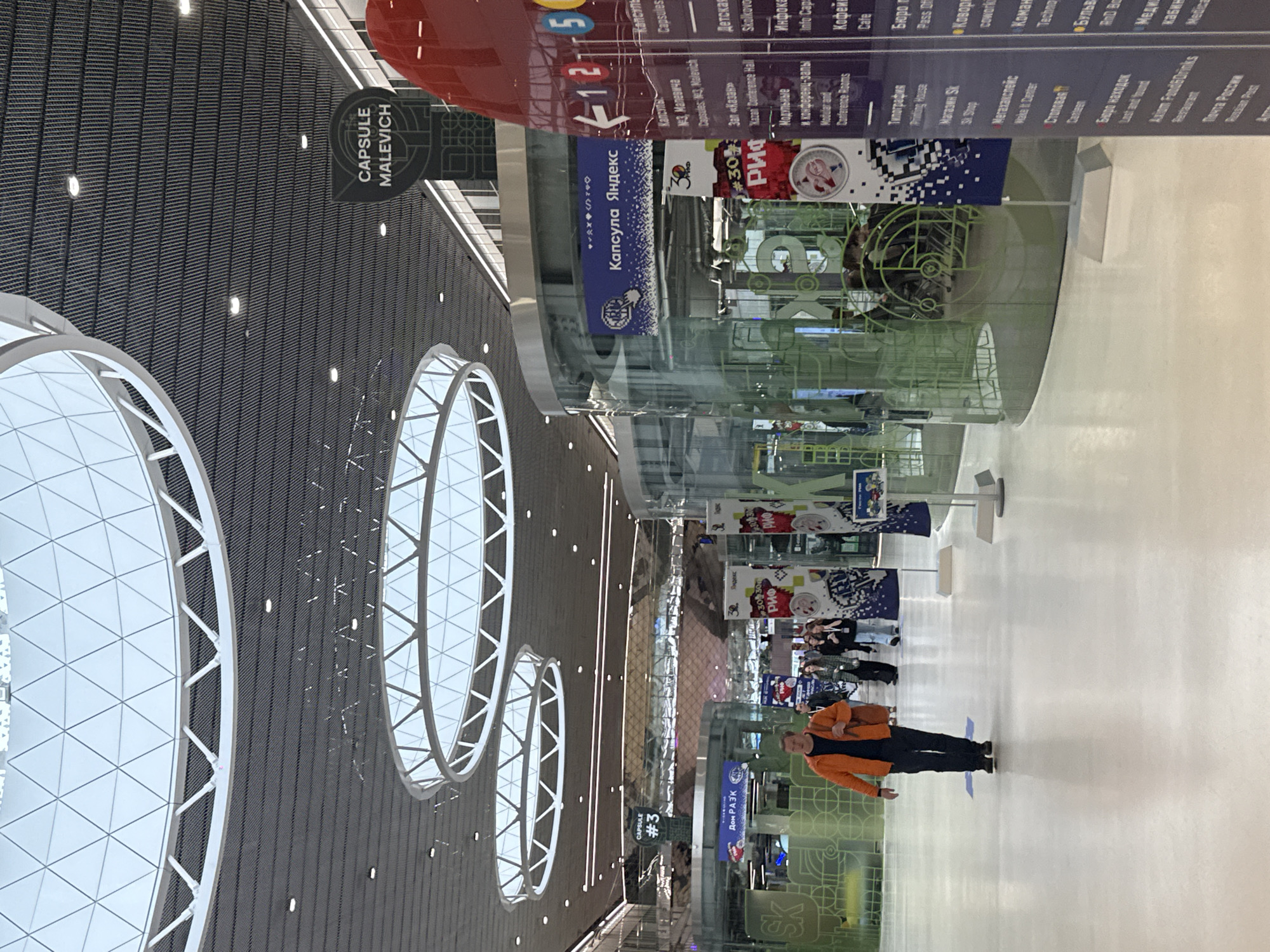 The image displays a brightly lit, modern indoor public space, likely a conference center, exhibition hall, or a large atrium, in Moscow, Russia. The ceiling features a dark, patterned surface with numerous embedded lights and several prominent, large, circular-to-oval skylight structures with intricate white framing.
In the midground, a man wearing an orange jacket and dark trousers stands facing right, looking towards the activity in the distance. Behind him and further down the hall, several other individuals are visible, walking or standing, suggesting a busy environment.
The space is characterized by glass partitions and tall information banners. Visible text, often in both Russian and English, indicates various facilities and services: "CAPSULE MALEVICH" and "Капсула Яндекс" (Yandex Capsule) point to dedicated pods or themed areas, possibly for work or privacy. Other signs list "Capsules: #3, Malevich," "Zal «Kazan»" (Kazan' Conference Hall), "Skolkovo," "Modular and Conference Rooms," "Info Cafe," "Printing House," "Sk Shop," "Wake It Center," "Canteen," "Cowork," "Food&Drinks," and "Russian Post." The presence of "РИФ" (RIF) and event-related signs suggests a forum or conference is taking place or has recently occurred.
A red cylindrical structure with numbers "125" and "3" is also visible, possibly indicating levels or zones. The overall atmosphere is professional and contemporary, typical of a modern innovation or business hub like Skolkovo, known for its focus on technology and startups. The lighting suggests daytime indoors.