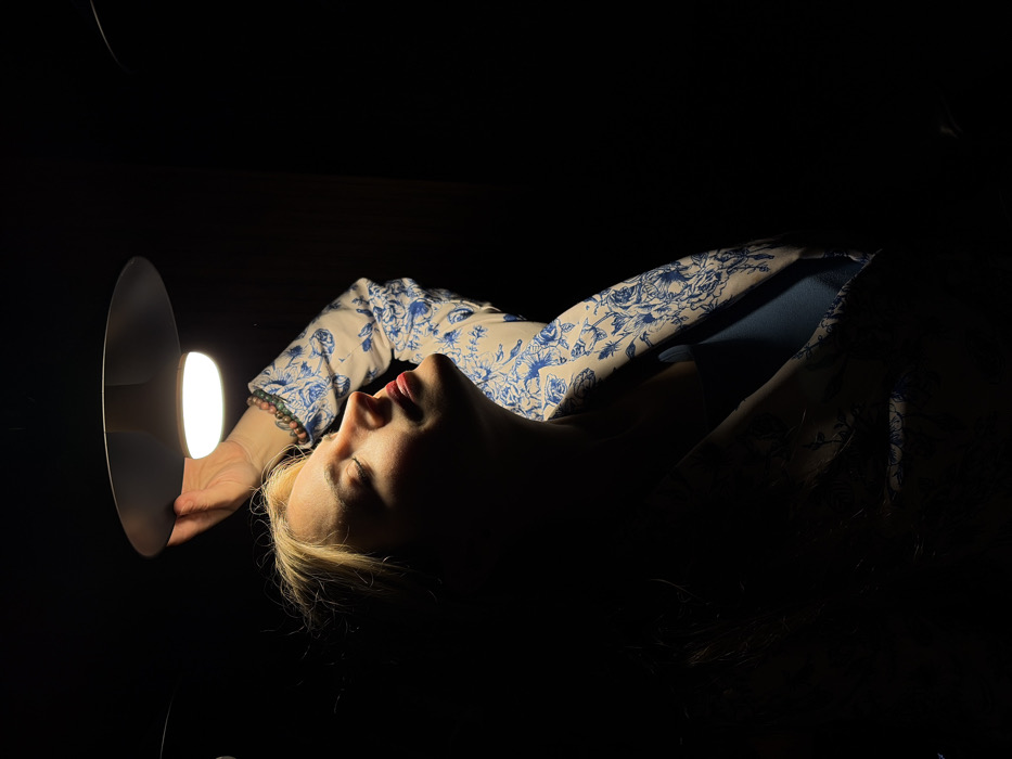 A woman lies illuminated by a lamp she holds, emphasizing her face, neck, and the blue floral pattern on her sleeve. Her eyes are closed, and her red lips are slightly parted. The surrounding space is dark, throwing her into stark relief. The darkness obscures the background and much of her figure, enhancing the dramatic lighting.