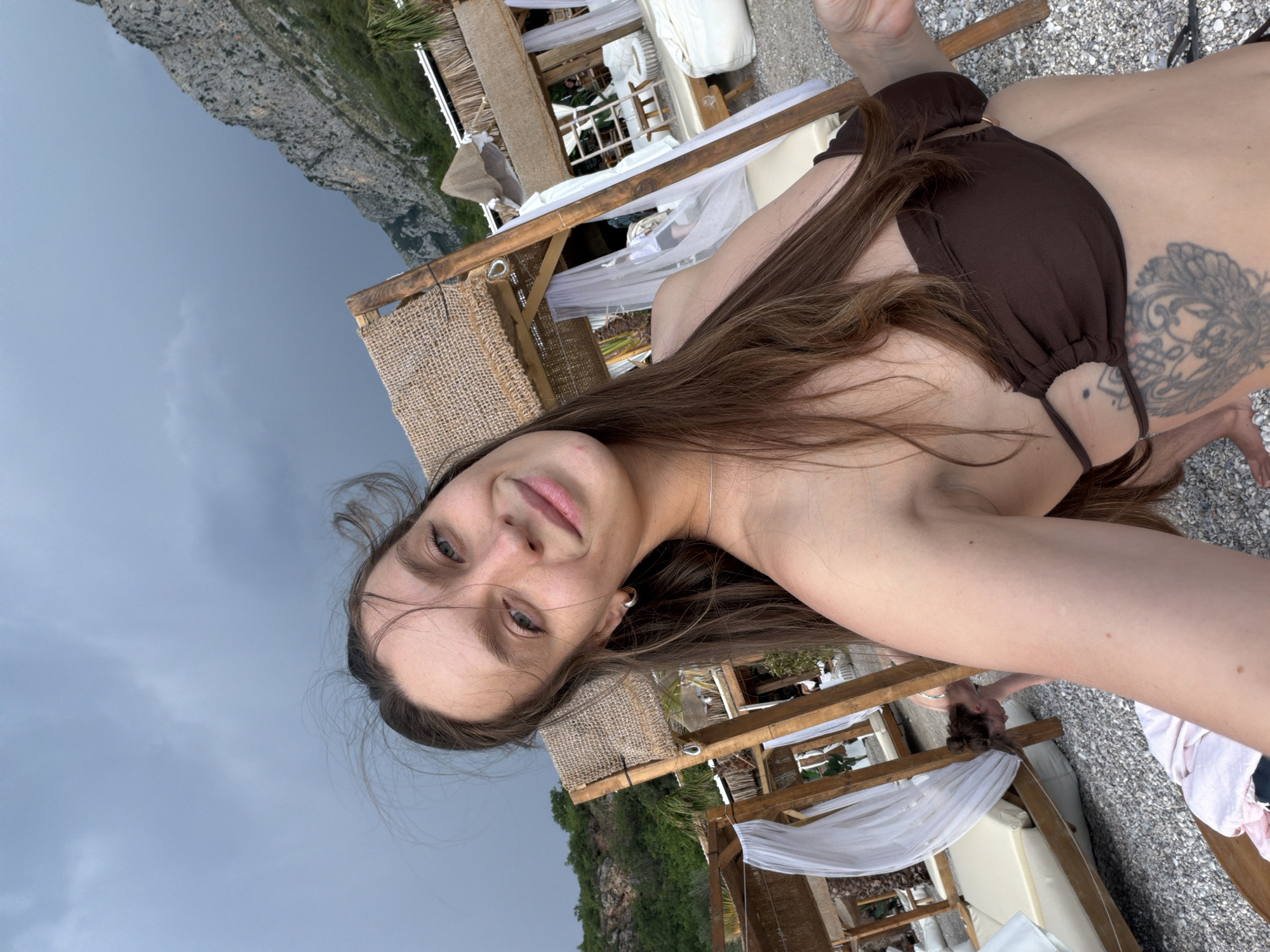 The media file shows a woman lying down on a beach, taking a selfie. She is wearing a brown bikini and has a tattoo on her side. The background includes beach chairs, umbrellas, and a rocky cliff face. The sky is overcast, suggesting it might be late afternoon or early evening. The overall atmosphere is relaxed and casual, consistent with a beach vacation. It is not possible to identify any specific text in the media file. The location is specified as Drobnići, Montenegro.