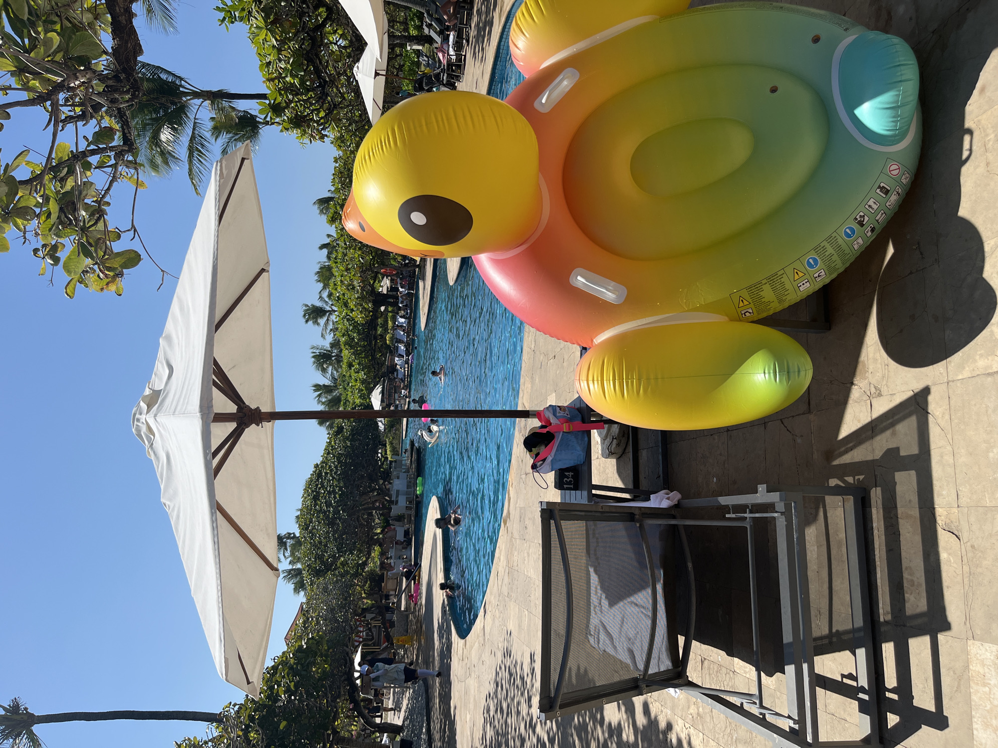 The image depicts a sunny day at a resort pool in Kuta Selatan, Indonesia. In the foreground, a grey lounge chair is positioned on a light-colored tiled patio. To the right of the chair is a large, colorful inflatable toy shaped like a cartoon character, featuring a yellow head with a brown eye and an orange and green body. Next to the inflatable toy is a small side table with a blue and pink bag and a white towel.
Beyond the foreground, a curved swimming pool with bright blue water is visible. Several people are in the pool, some swimming and others relaxing on inflatable rings. Lush green trees and palm trees surround the pool area, with white umbrellas providing shade. In the background, resort buildings and more trees can be seen under a clear blue sky. The overall atmosphere is one of leisure and vacation.