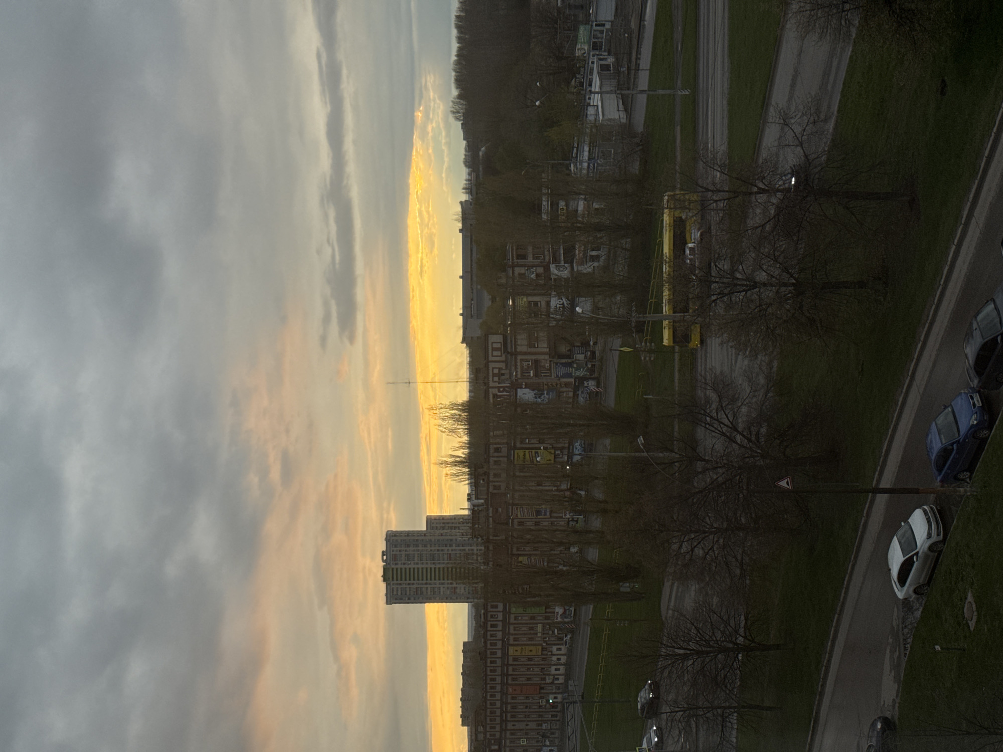 The image depicts an urban landscape at either dawn or dusk, viewed from an elevated perspective, possibly a window of a building. The sky dominates the upper portion, featuring dense grey clouds that transition to a vibrant band of orange and yellow light along the horizon on the left.
In the mid-ground, a series of multi-story residential or commercial buildings are visible, characterized by their blocky architecture. One prominent tall, light-colored high-rise building stands out against the glowing horizon. Many bare-branched trees are scattered throughout the scene, suggesting a late autumn, winter, or early spring season.
Below the buildings, a network of roads and paved areas runs through grassy verges. Several cars are parked along the roads, including a white sedan, a blue car, and other darker vehicles. A distinctive yellow bus or commercial vehicle is also visible in the mid-ground. Streetlights and utility poles are sparsely placed.
No human activity is directly discernible, conveying a sense of quiet urban life. The overall lighting indicates a transitional time of day, with the soft, warm light from the horizon contrasting with the muted tones of the buildings and the dark, cloudy sky. A red triangular "Yield" sign with a white background is visible on a pole near the road. Text on banners attached to some buildings is present but illegible due to blurriness.
