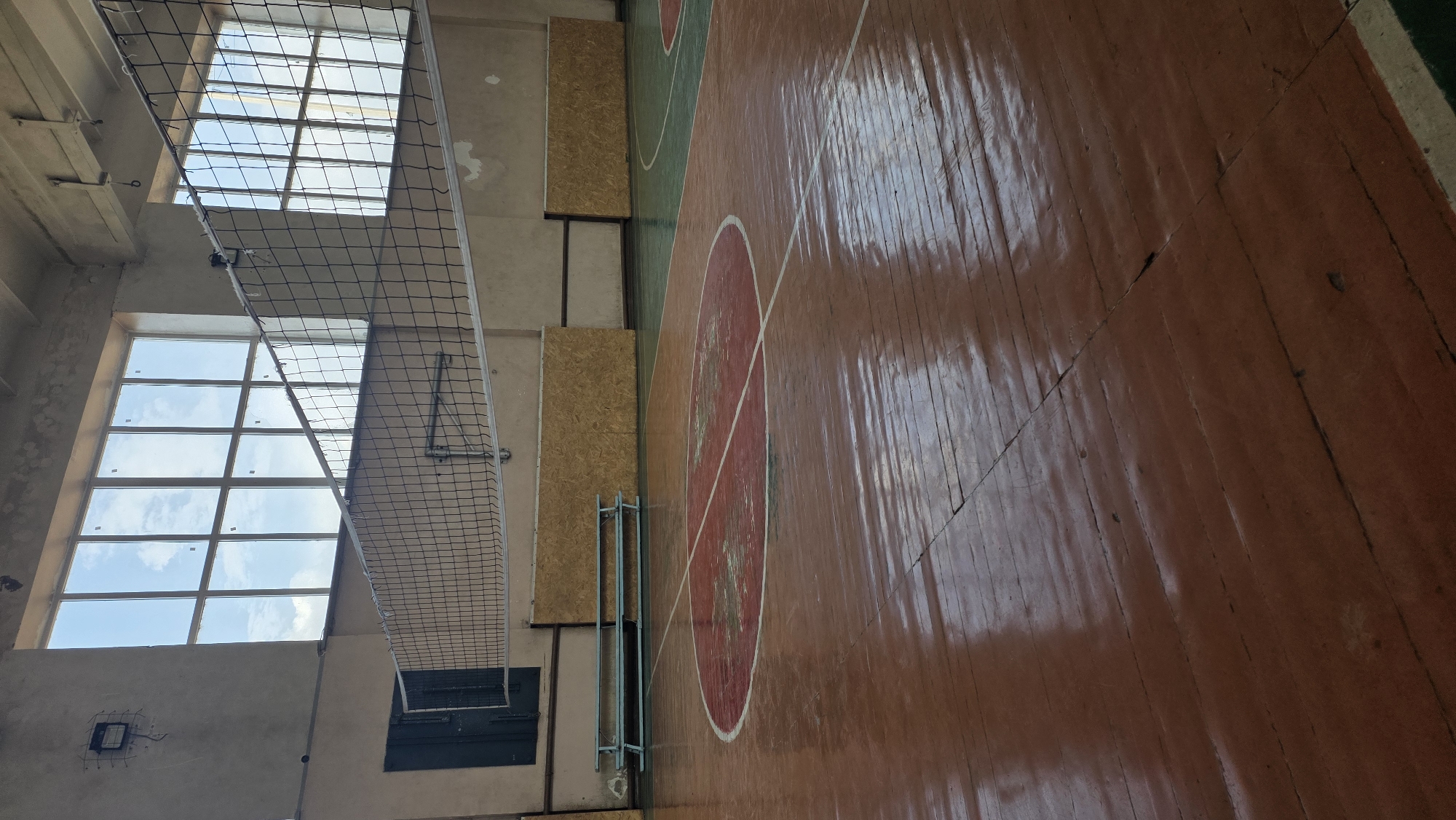 The image shows an interior view of a volleyball court in Tokmok City, Kyrgyzstan. The court features a polished wooden floor with red and white markings. A volleyball net is suspended across the middle of the court. In the background, there are large windows looking out to a cloudy sky, and along the walls, there are benches and wooden panels. The overall impression is of an indoor sports facility, likely used for physical education or sports events.
