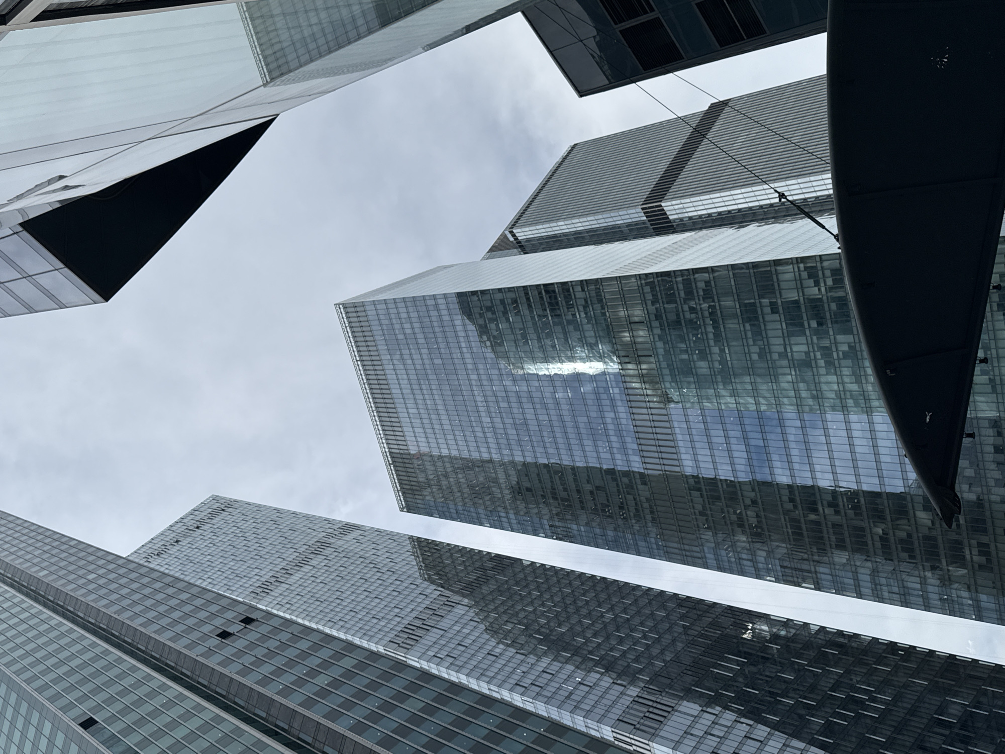 The image shows a low-angle view of modern glass skyscrapers. The sky is overcast, creating a diffuse lighting across the scene. The architectural style is contemporary, with reflective glass facades. No people are visible. There is a black object on the top right of the image. It is of unknown origin. The location is specified as Khimki, Russia.