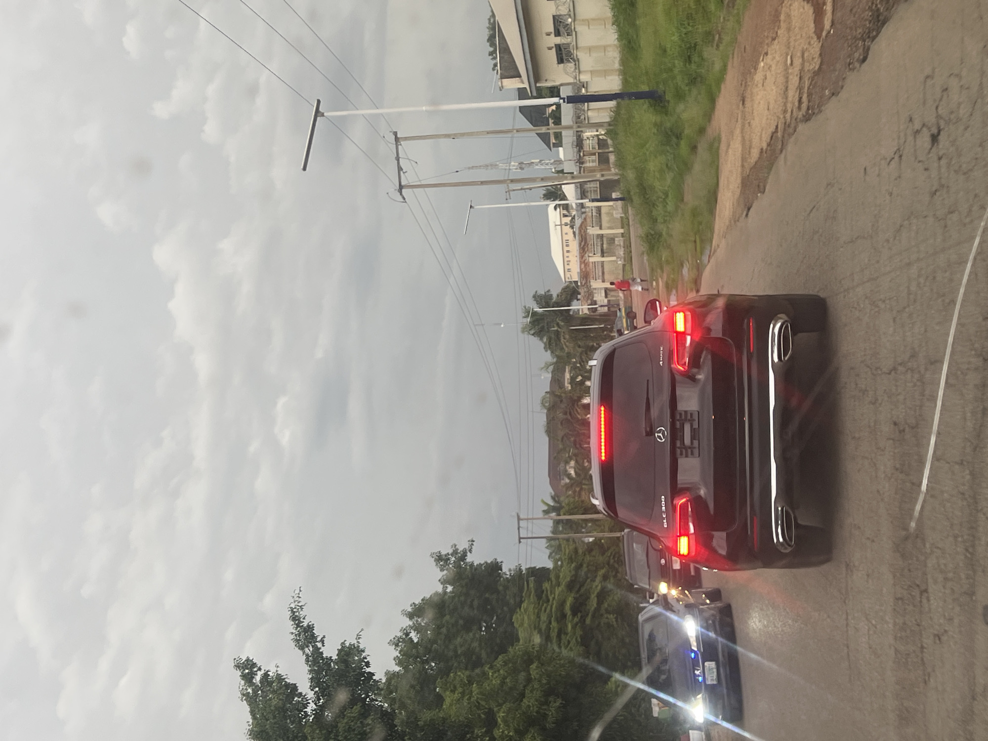 The image shows a road scene in Jos, Nigeria, with several cars driving on a paved road. The main focus is on the rear of a dark-colored Mercedes-Benz GLC 300, with its brake lights illuminated. Behind it, another dark-colored vehicle is visible, followed by a lighter-colored car with its headlights on. The road appears to be in a somewhat degraded condition, with visible cracks.
The scene is set in an outdoor environment with a cloudy sky, suggesting overcast weather or perhaps late afternoon. On the left side of the road, there are tall utility poles with electrical wires, and further back, buildings are visible, some appearing to be commercial or industrial structures with barbed wire on top. Lush green vegetation, including trees and grass, lines the right side of the road. A few individuals are seen walking on the side of the road in the distance, near the buildings.
The overall impression is that of a typical day with traffic on a road in a developing area, possibly on the outskirts of a town or city, given the mix of infrastructure and vegetation. There are no strong emotional cues conveyed by the image. The visible text on the car is "GLC 300".