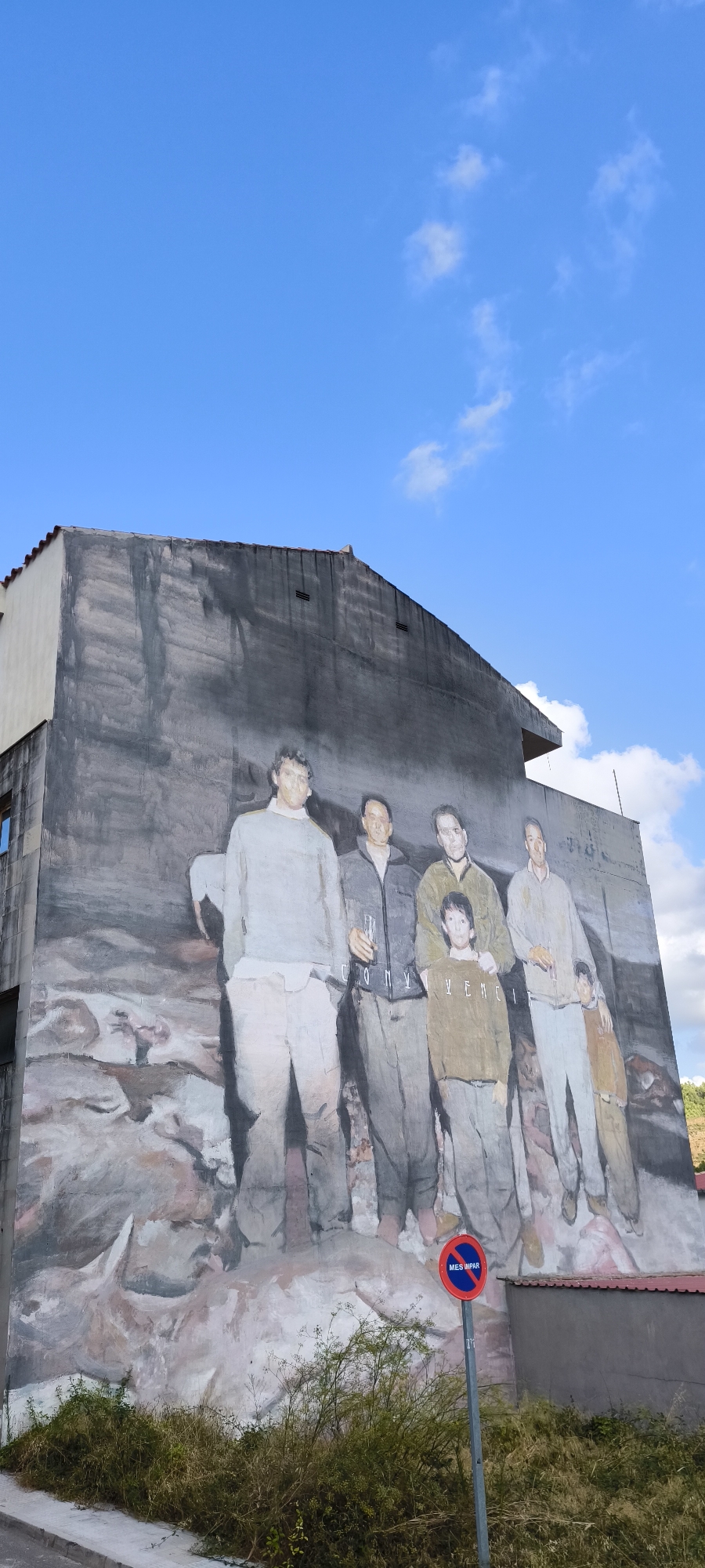 The image depicts a large mural on the side of a building in Fanzara, Spain. The mural is a lifelike painting of a group of six people, appearing to be men and boys, standing together in what seems to be a natural or rugged landscape. From left to right, the first person is a tall man in a light grey sweatshirt and light-colored pants. The second is a man in a dark jacket, holding a glass. The third is a man in a dark green jacket, with his arm around a young boy in front of him who is wearing a brown sweatshirt. The fifth person is a man in a light grey shirt and pants, and the sixth is a young boy in a brown jacket, standing to the right of the fifth person. The mural covers a significant portion of the building's wall, which is painted in muted, earthy tones. The building itself is a multi-story structure with a pitched roof. In the foreground, there is dense green vegetation and a sidewalk. A blue and red circular sign with the words "MES IMPAR" is visible on a pole in front of the mural, indicating parking restrictions for odd-numbered days. The sky above is a clear blue with scattered white clouds.