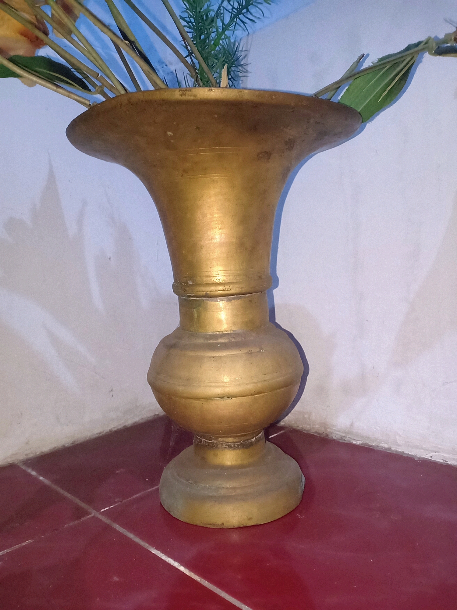 A brass vase with a wide, flaring rim sits on a dark red tiled surface in the corner of a room. The vase has a bulbous body and a flared base. Several dried plant stems and green leaves are arranged inside the vase. The background consists of a white wall with subtle shadows. The image appears to have been taken indoors, possibly in a residential setting in Jarit, Indonesia. There are no people or animals visible in the scene.