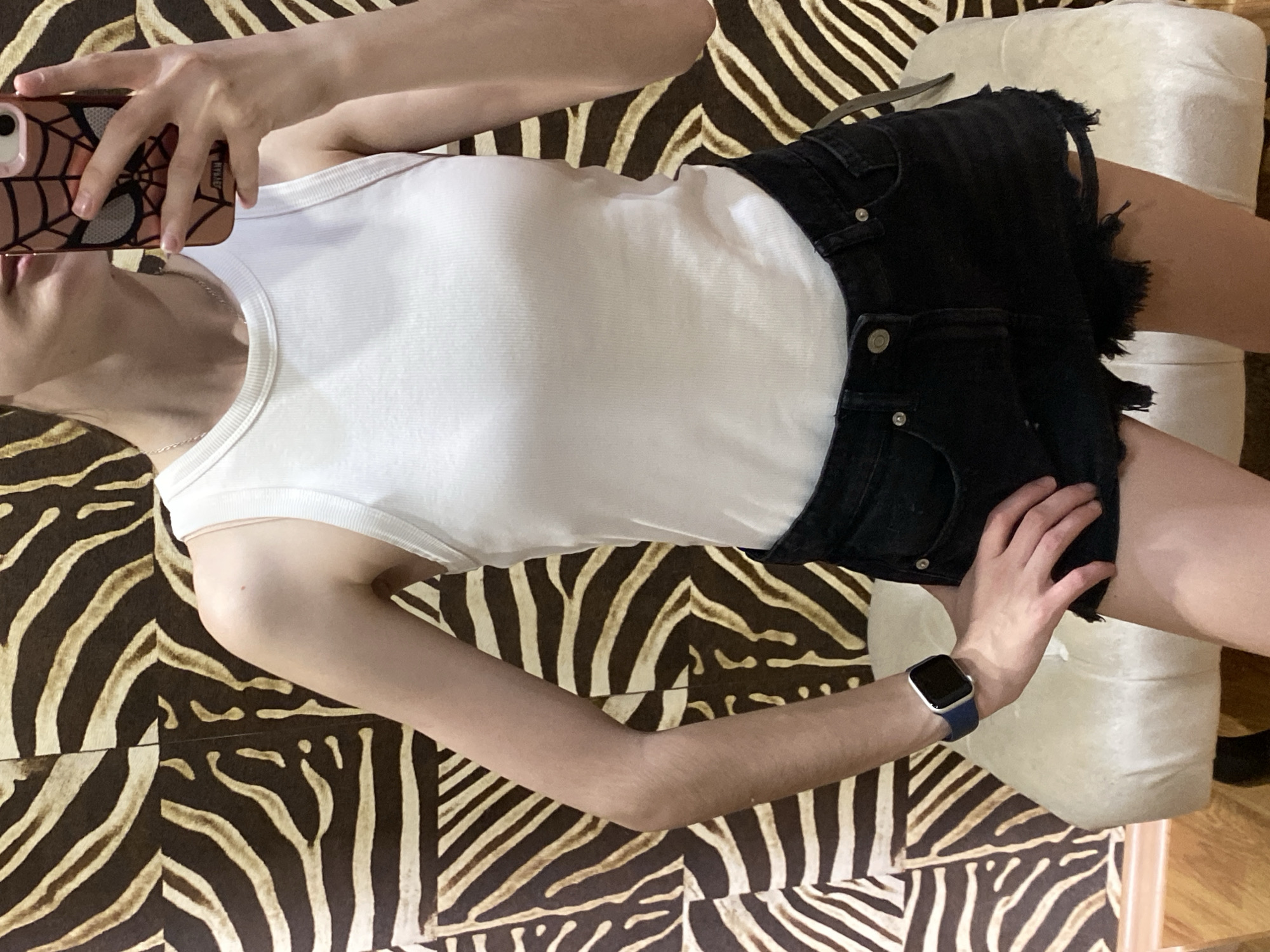 A person wearing a white tank top and black denim shorts lies on a zebra-print upholstered surface. The person is holding a smartphone with a Spider-Man-themed case to take a selfie. The left hand is resting on the denim shorts. A smartwatch with a blue band is visible on the left wrist. A white cushion is present in the background. The location is not specified in the city of Votkinsk, Russia.